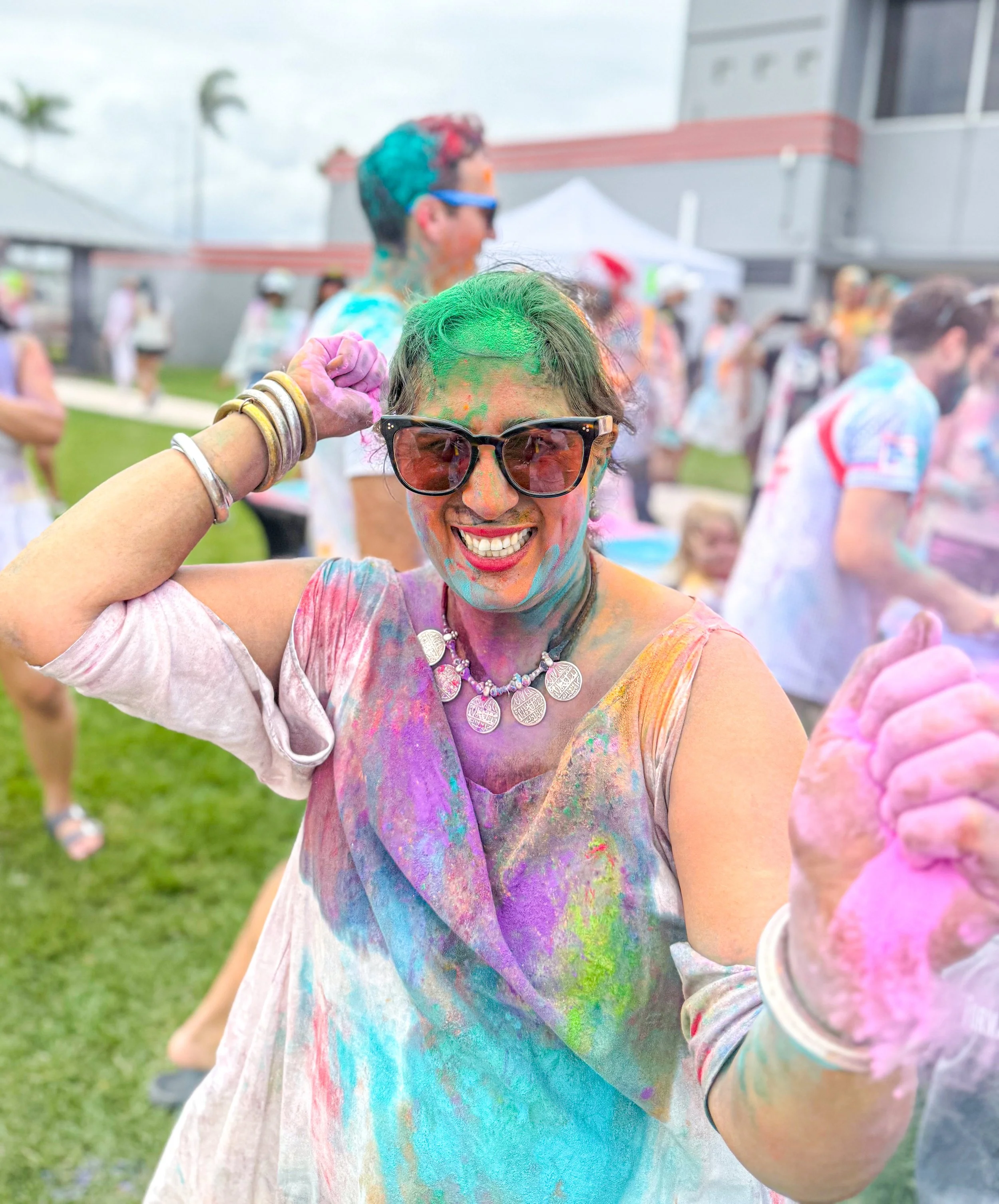 Woman smiling and wearing sunglasses, covered in colorful powder at a festival.