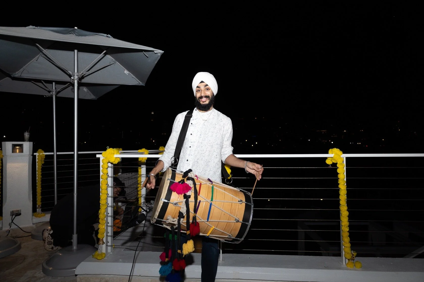 A man with a white turban and beard smiling, playing a drum on a balcony at night with city lights in the background, decorated with yellow marigold flowers.
