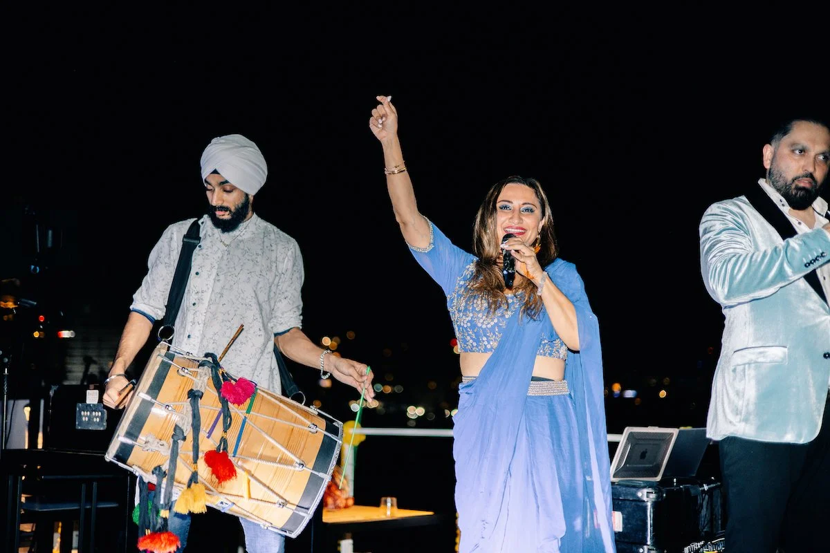 A woman in a blue sari singing into a microphone on stage, with a man playing a drum and another man with a microphone standing nearby during an outdoor night event.