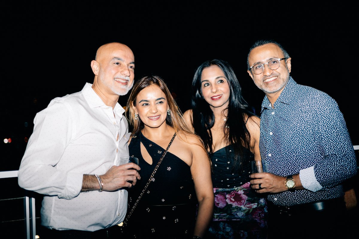 Four adults standing close together at a nighttime event, smiling at the camera, with some holding drinks.