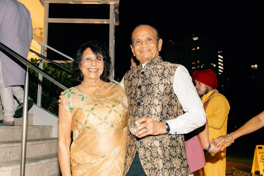 Indian couple at night, the woman in a gold saree with floral patterns and the man in a sherwani with floral designs, smiling at an outdoor event.