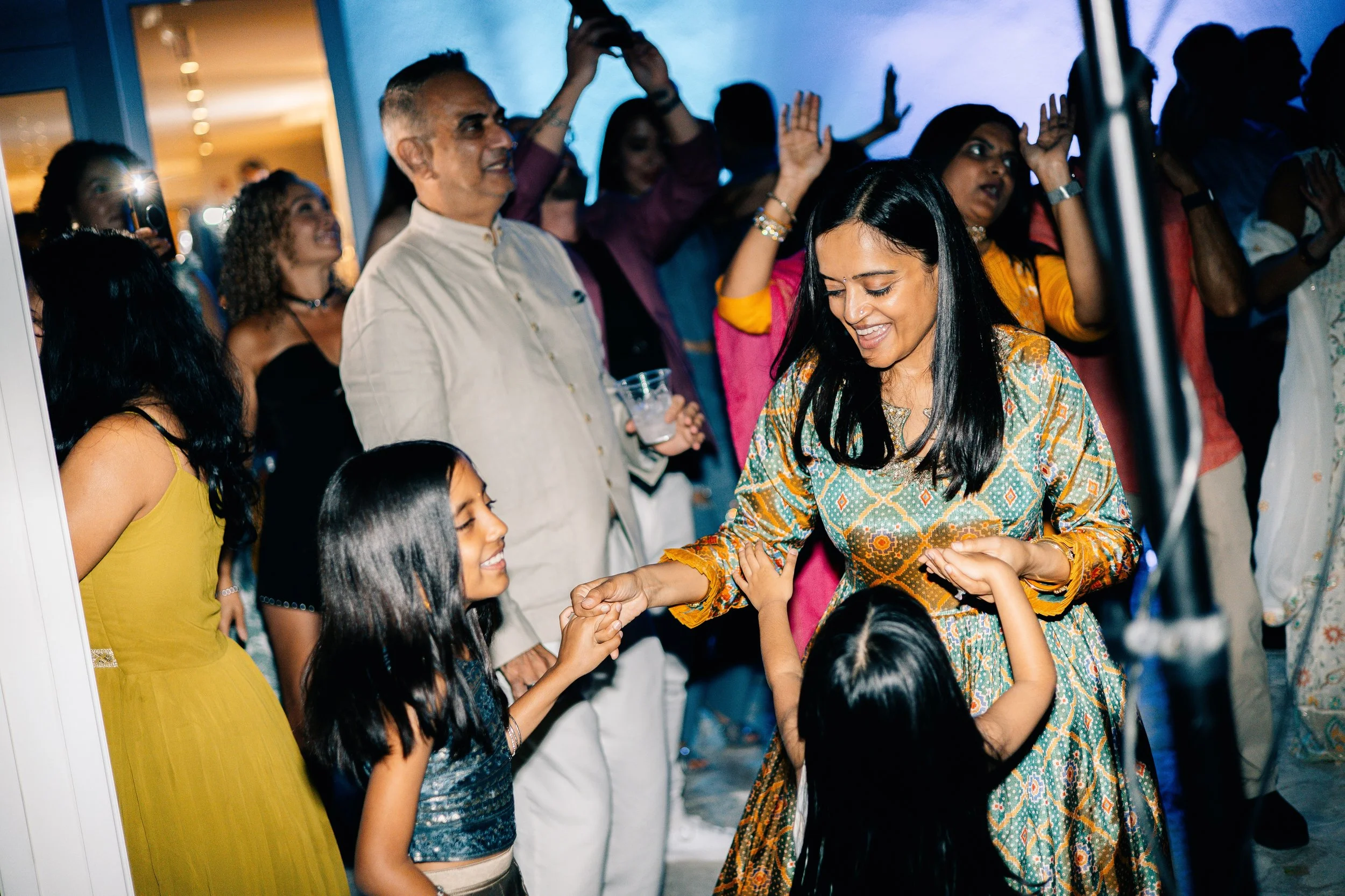 A woman in a patterned dress dancing and smiling with two young girls, surrounded by other people at a lively gathering or celebration.