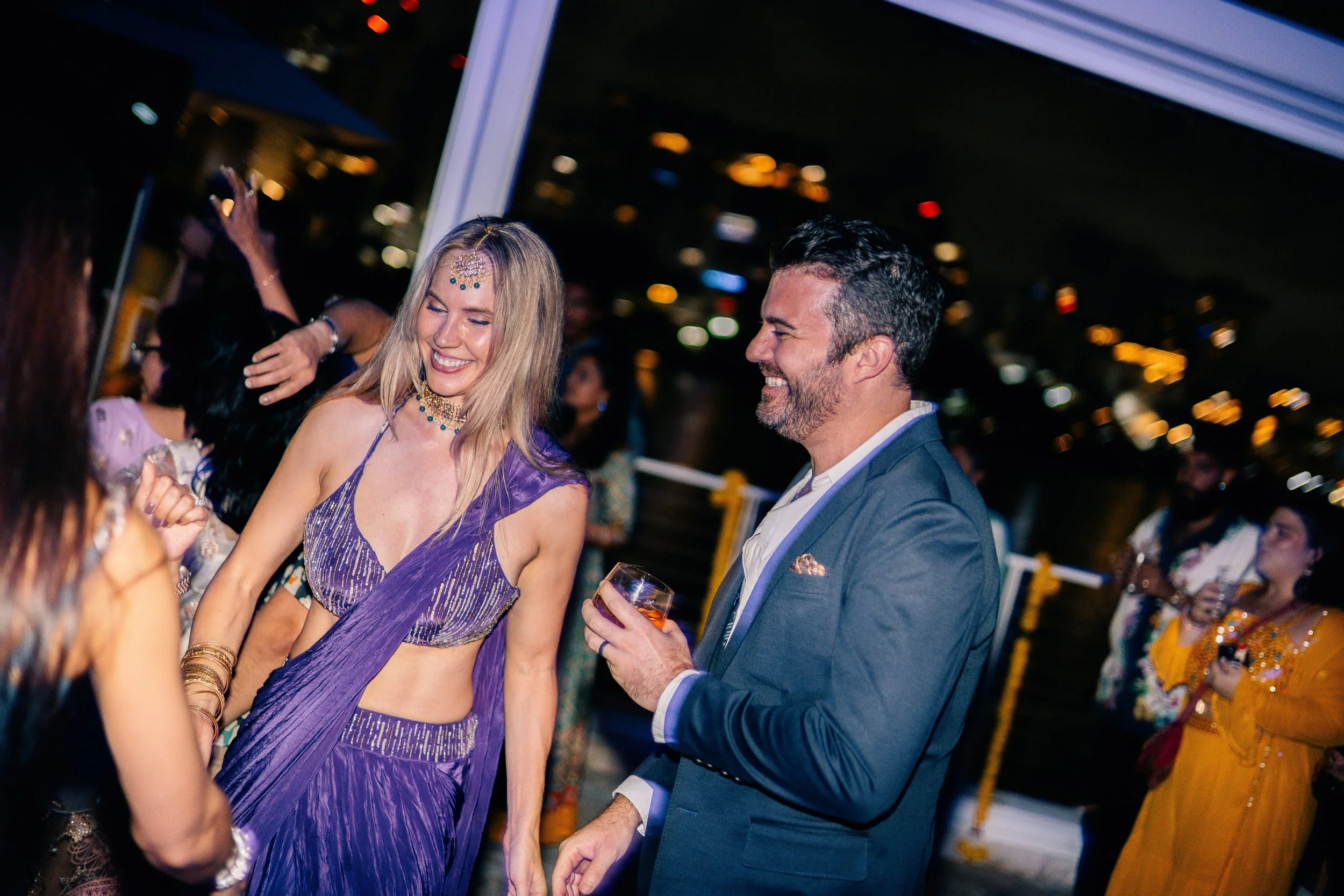 A woman and a man at a party, laughing and smiling. The woman wears a purple traditional dress with jewelry, and the man wears a blue suit, holding a drink.