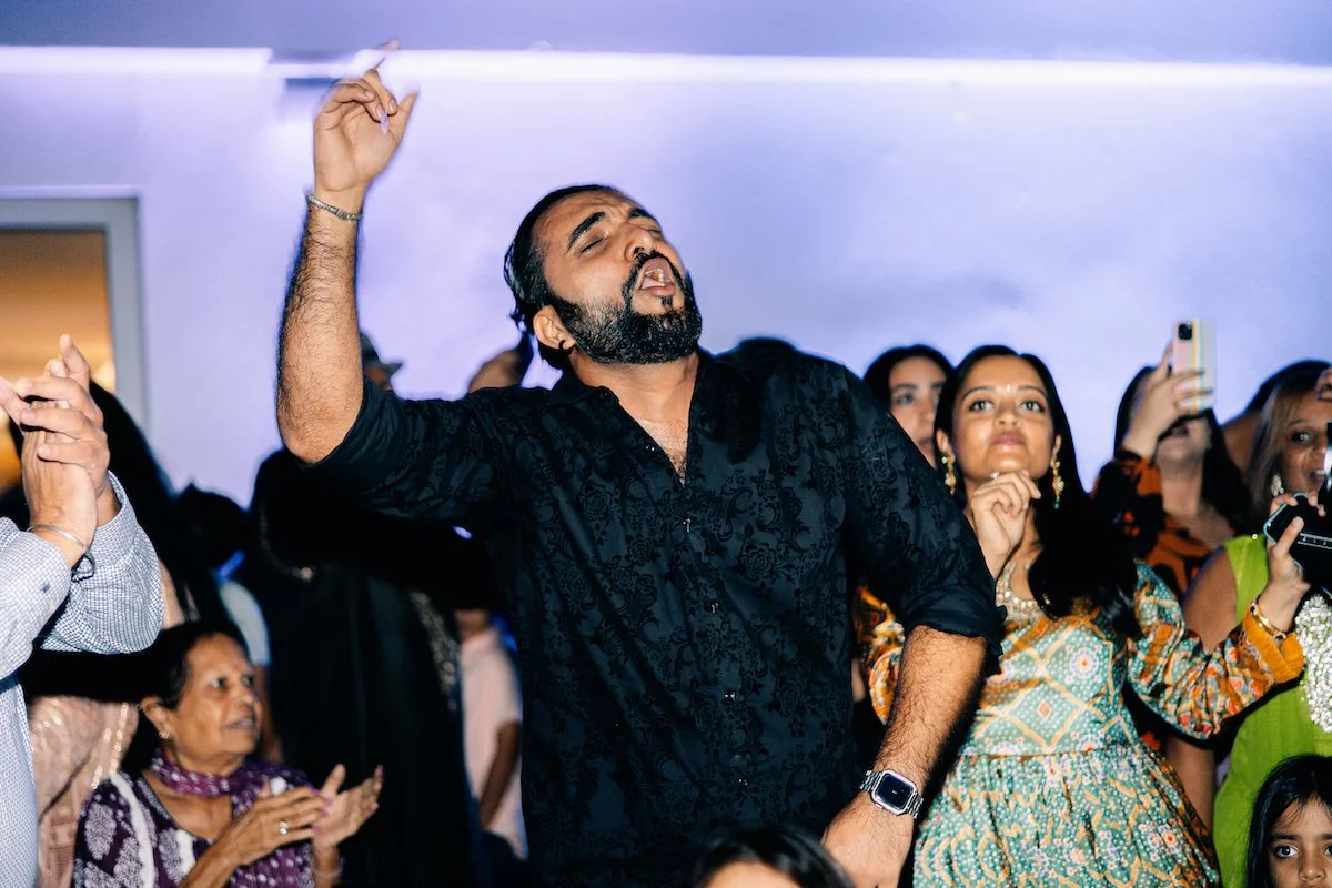 A man dancing passionately with eyes closed and his right arm raised in a crowded indoor party or celebration, surrounded by women and other guests watching and taking photos.