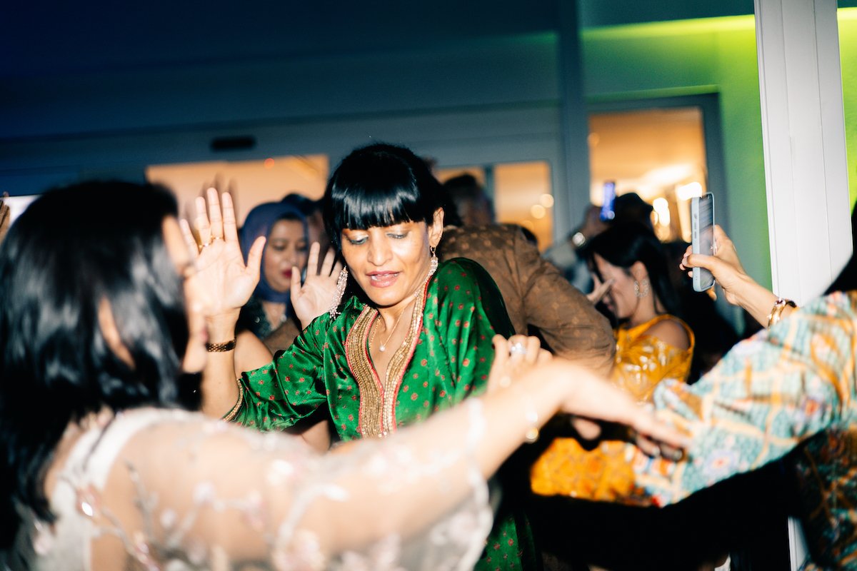 A group of women dancing and celebrating at a party or wedding reception. The woman in focus is wearing a green traditional dress with gold accents, and others are dressed in elegant attire.