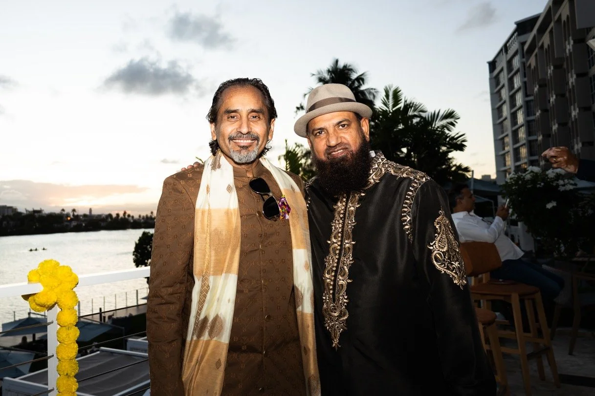 Two men dressed in traditional Indian attire standing outdoors during sunset with a river and cityscape in the background.