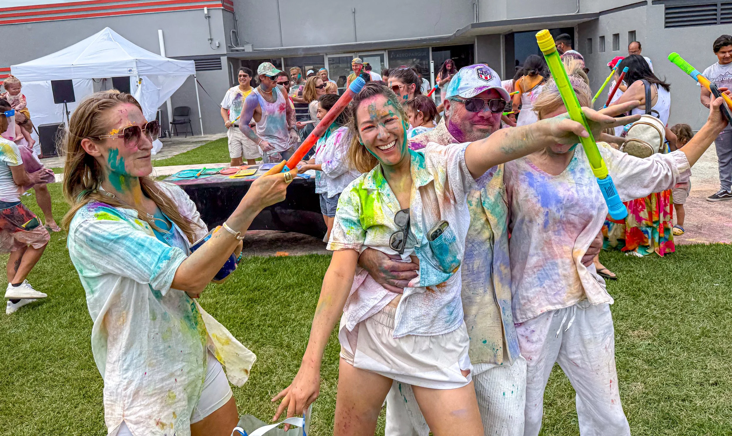 Group of people celebrating at a colorful Holi festival, throwing colored powders and holding water guns, with smiles and laughter on their faces.