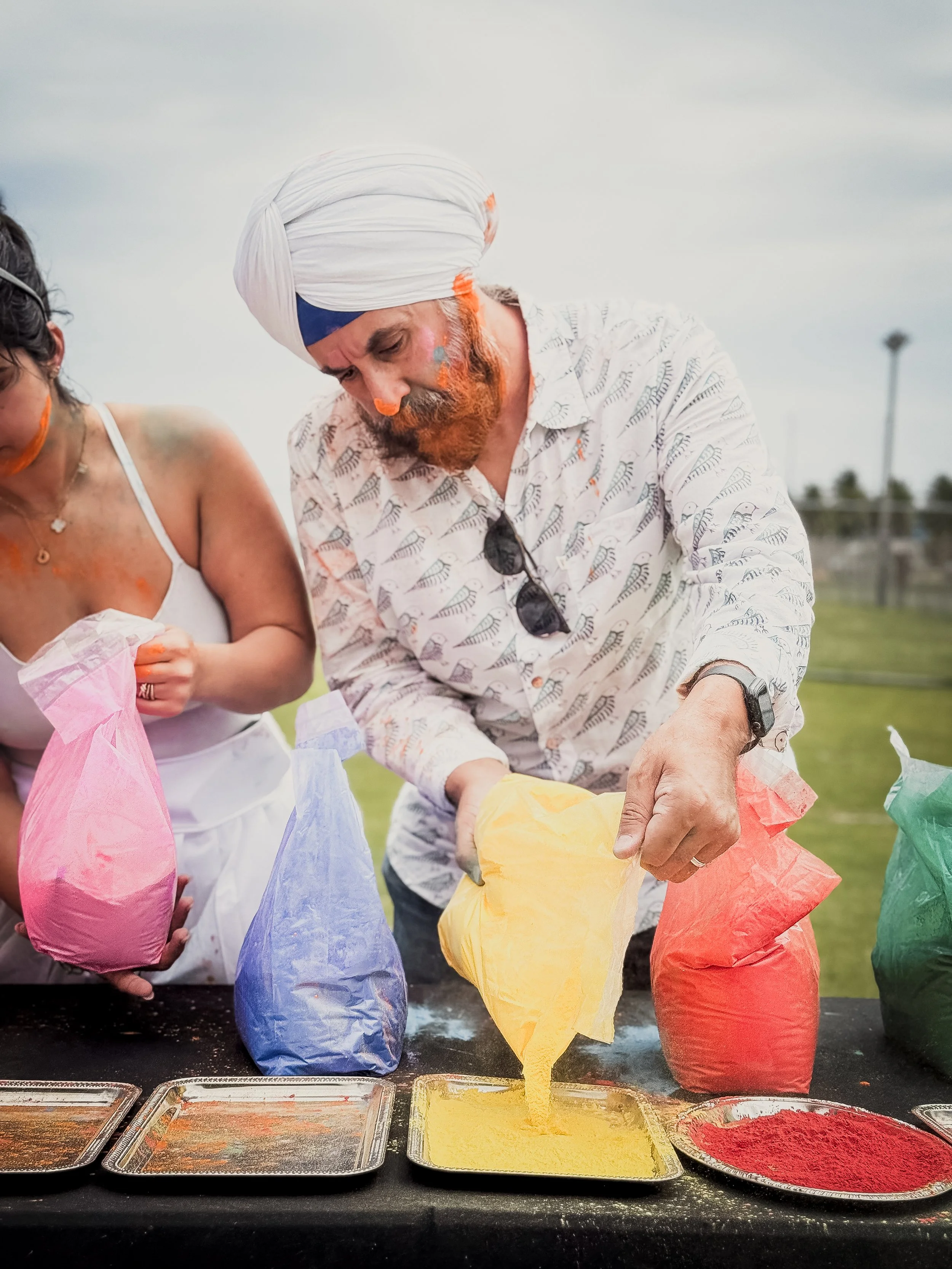 Man and woman participating in a colorful powder throwing event, filling paper bags with colored powder, outdoors on a cloudy day.