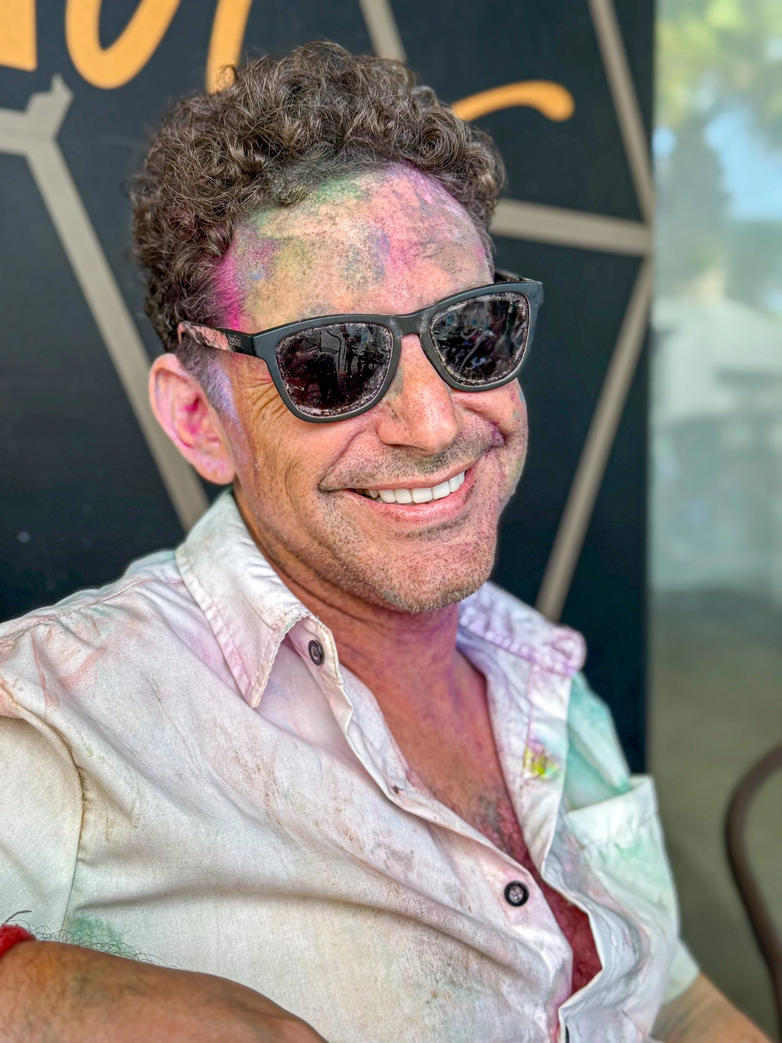 Man smiling wearing sunglasses with colorful chalk or powder on his face, in a casual shirt, at a festive outdoor event.