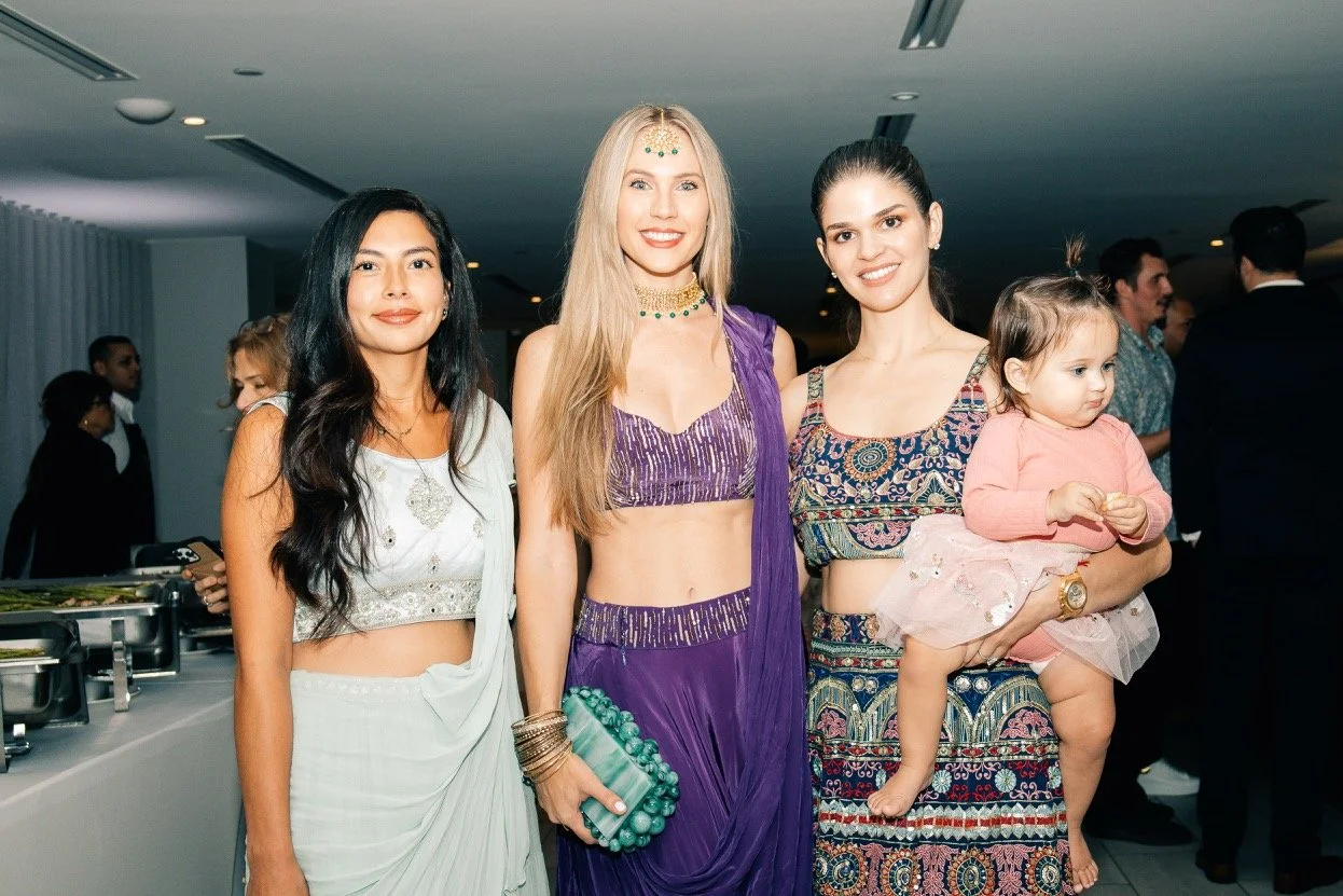 Four women, one holding a young girl, stand together at a social gathering, dressed in colorful traditional and modern outfits.