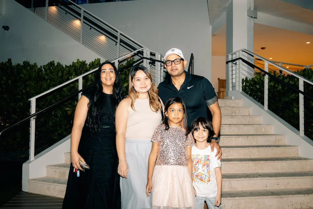 Group of five people, three women and two children, standing on a staircase outdoors at night. The woman on the left is wearing a black dress, the woman in the middle is wearing a beige top and a white skirt, the man is wearing glasses, a black shirt
