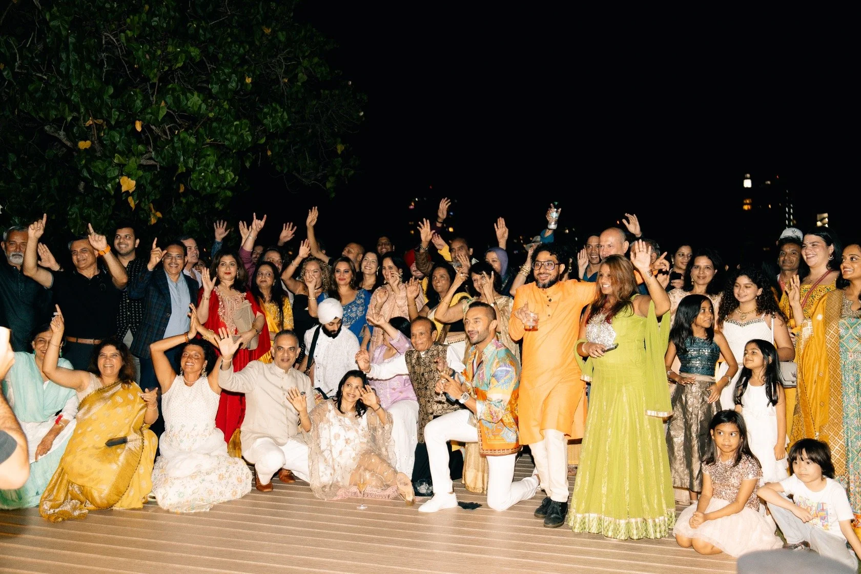 A large group of people celebrating outdoors at night, smiling and raising their hands, with some seated and others standing.