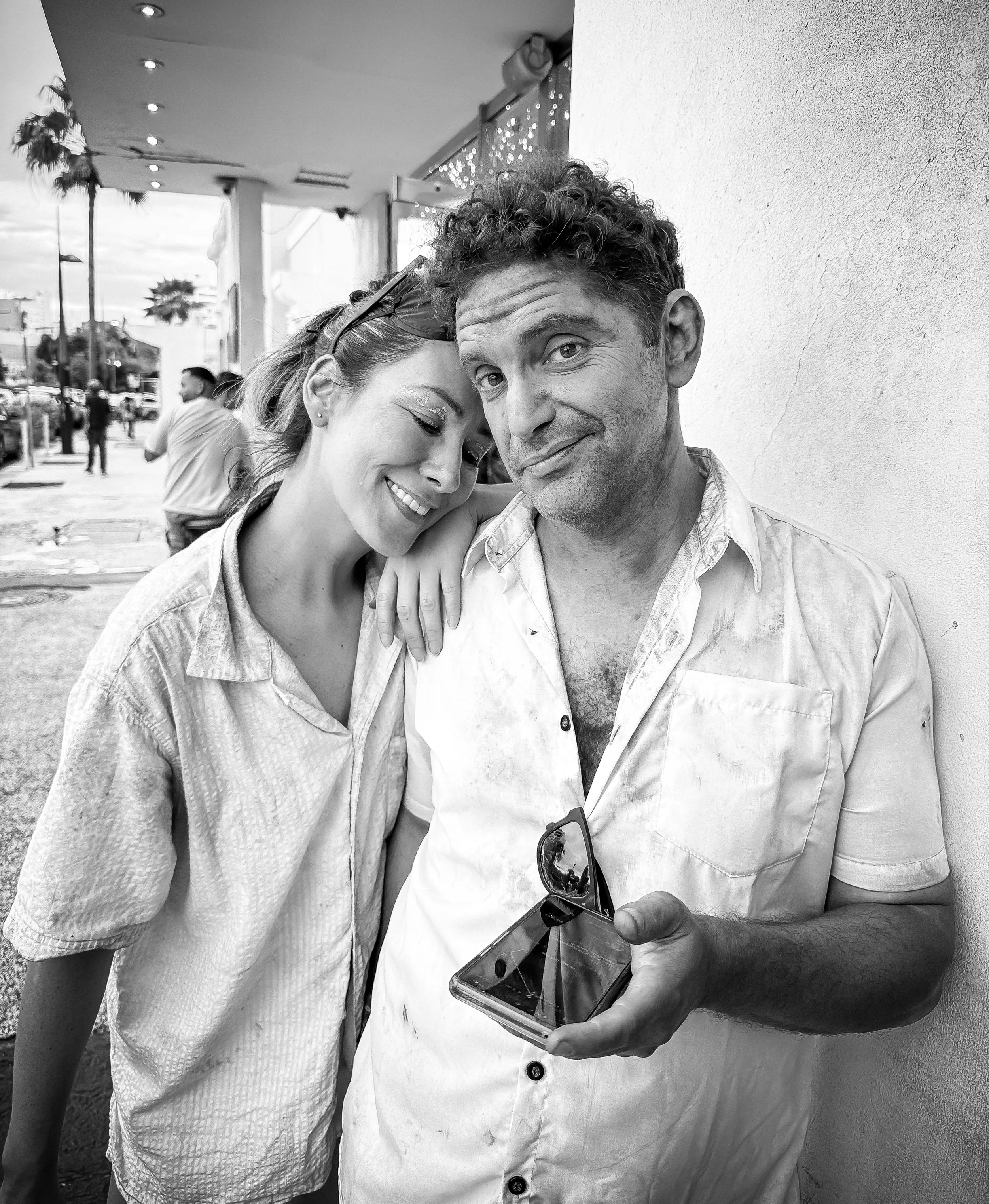 A man and woman stand close together outside, smiling. The woman leans on the man's shoulder, and the man holds sunglasses and a smartphone. It is a black and white image.