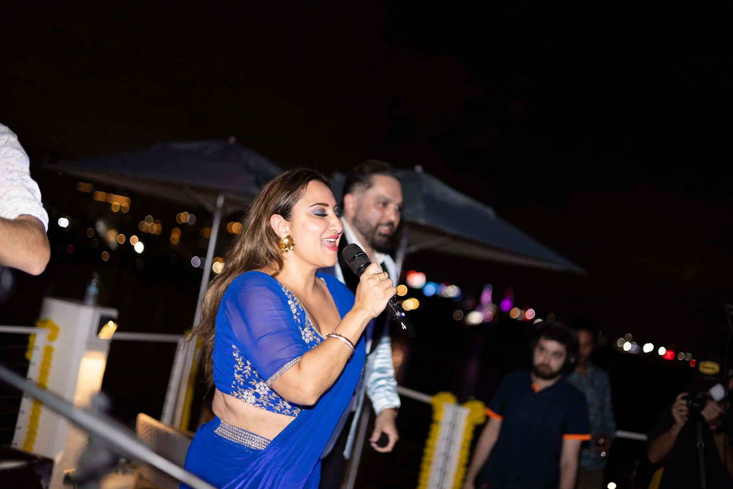 A woman in a royal blue sari with gold embellishments, holding a microphone and smiling, during an outdoor night event with a man in a light-colored shirt behind her and blurred city lights in the background.