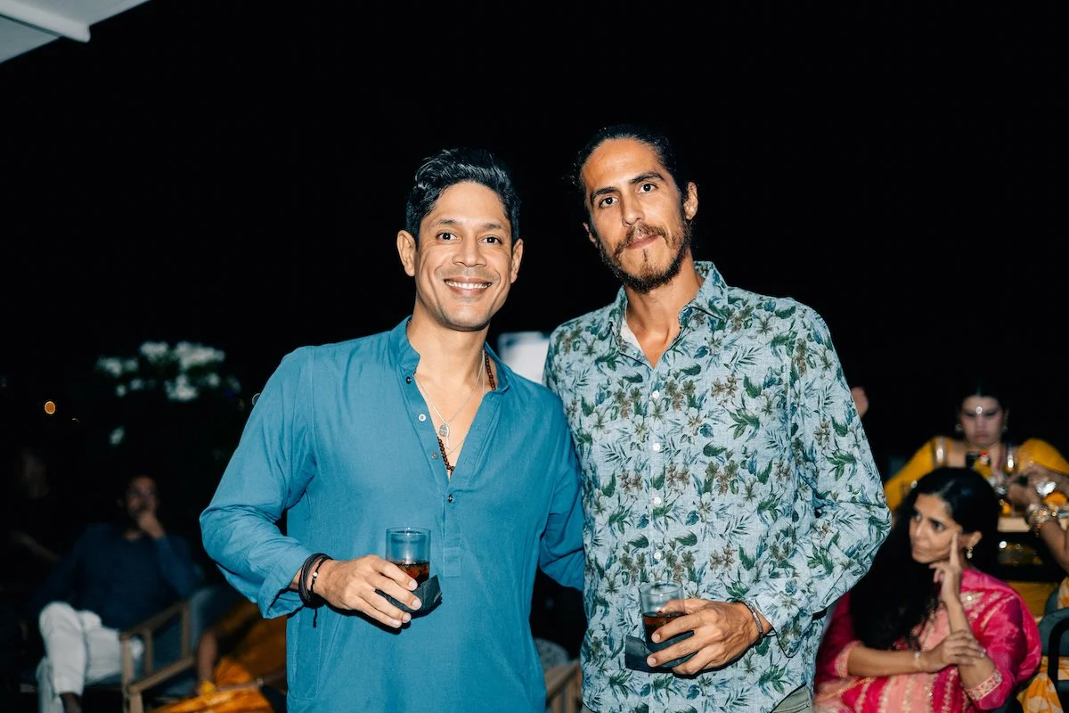 Two men smiling at a nighttime outdoor event, holding drinks, with other guests in the background.