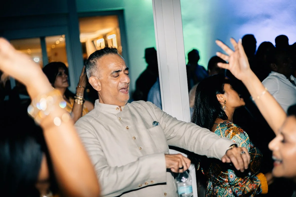 A man with short hair and a light-colored traditional shirt is dancing with his eyes closed and an expression of enjoyment at a social gathering. Surrounding people are also dancing and celebrating.