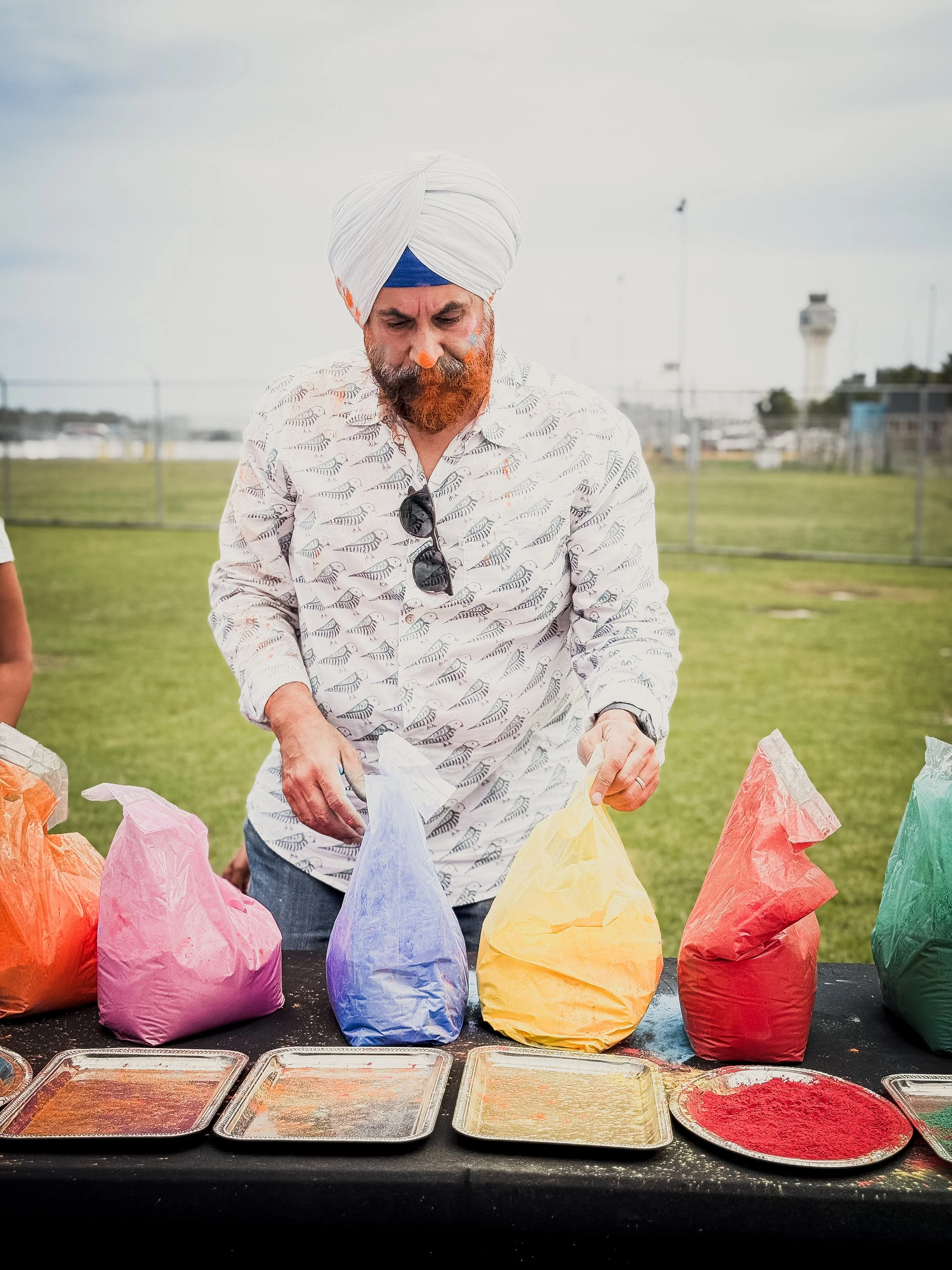 A man wearing a turban and patterned shirt is standing behind a table with colorful paper bags and trays of colored powders, likely preparing for a celebration or festival outdoors.