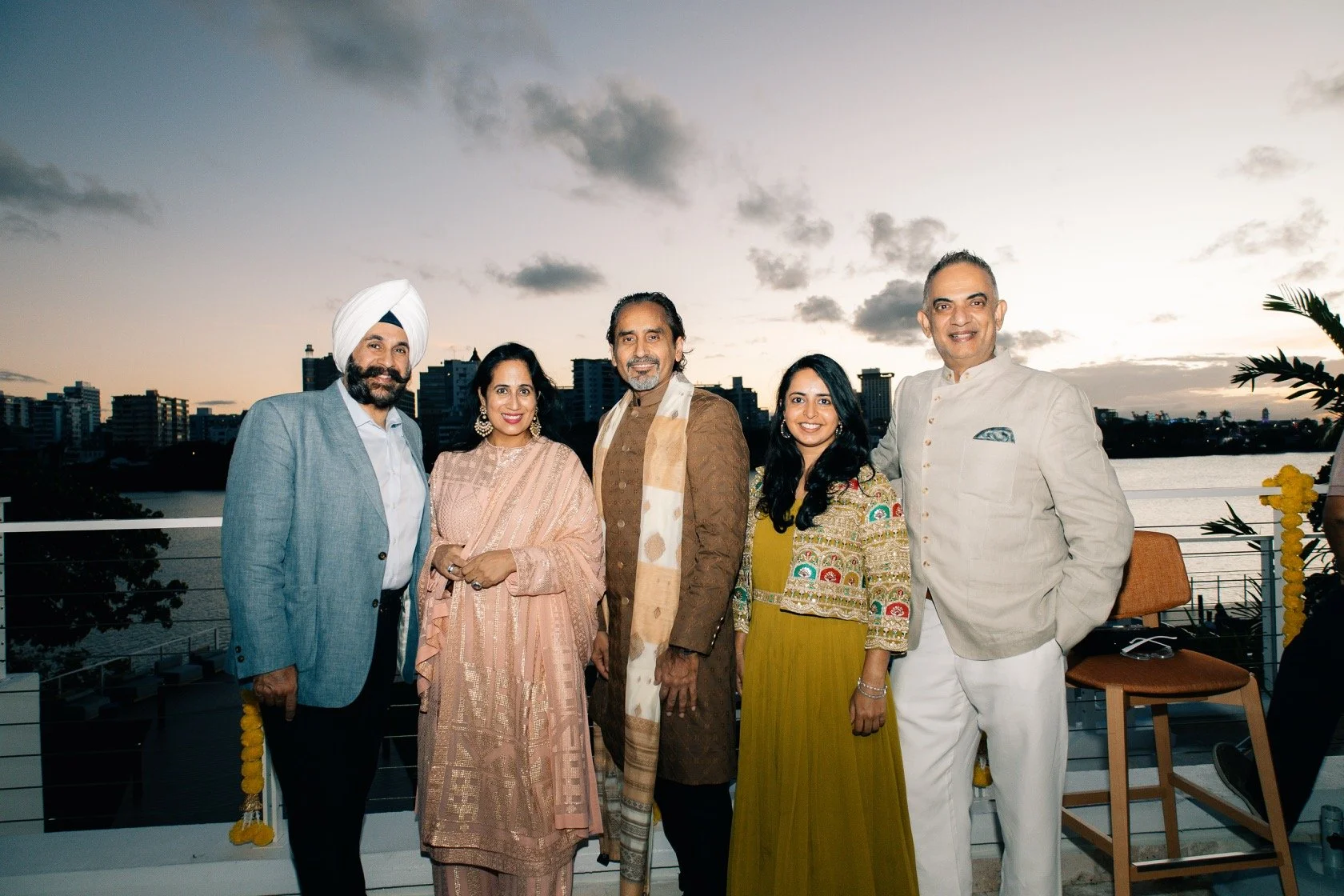 Group of five people dressed in traditional Indian attire at a social gathering on a rooftop during sunset, with city skyline and river in background.
