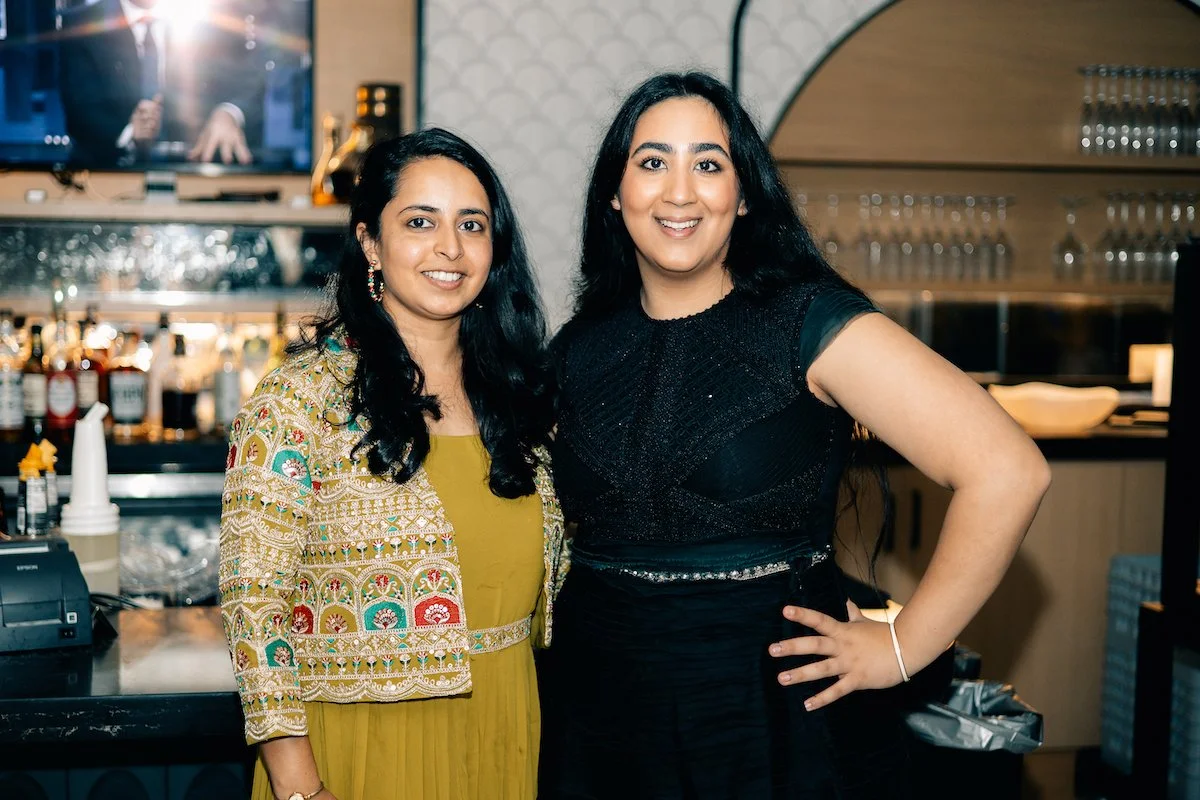 Two women smiling and standing in a bar or restaurant, with a counter and bottles of alcohol in the background.