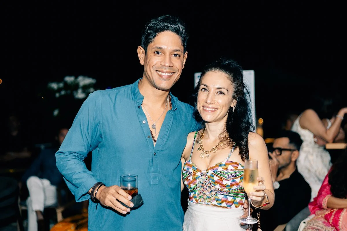 A smiling man and woman at an evening event, holding drinks, with other guests seated behind them.