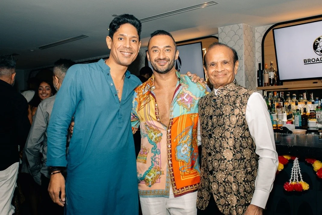 Three men posing together at a social event with bar and drinks in the background.