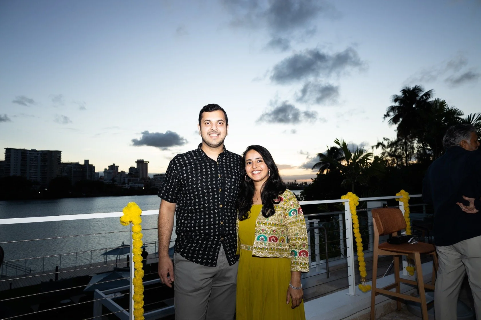 A couple standing on a balcony during sunset, with city buildings, water, and palm trees in the background, decorated with yellow flowers.