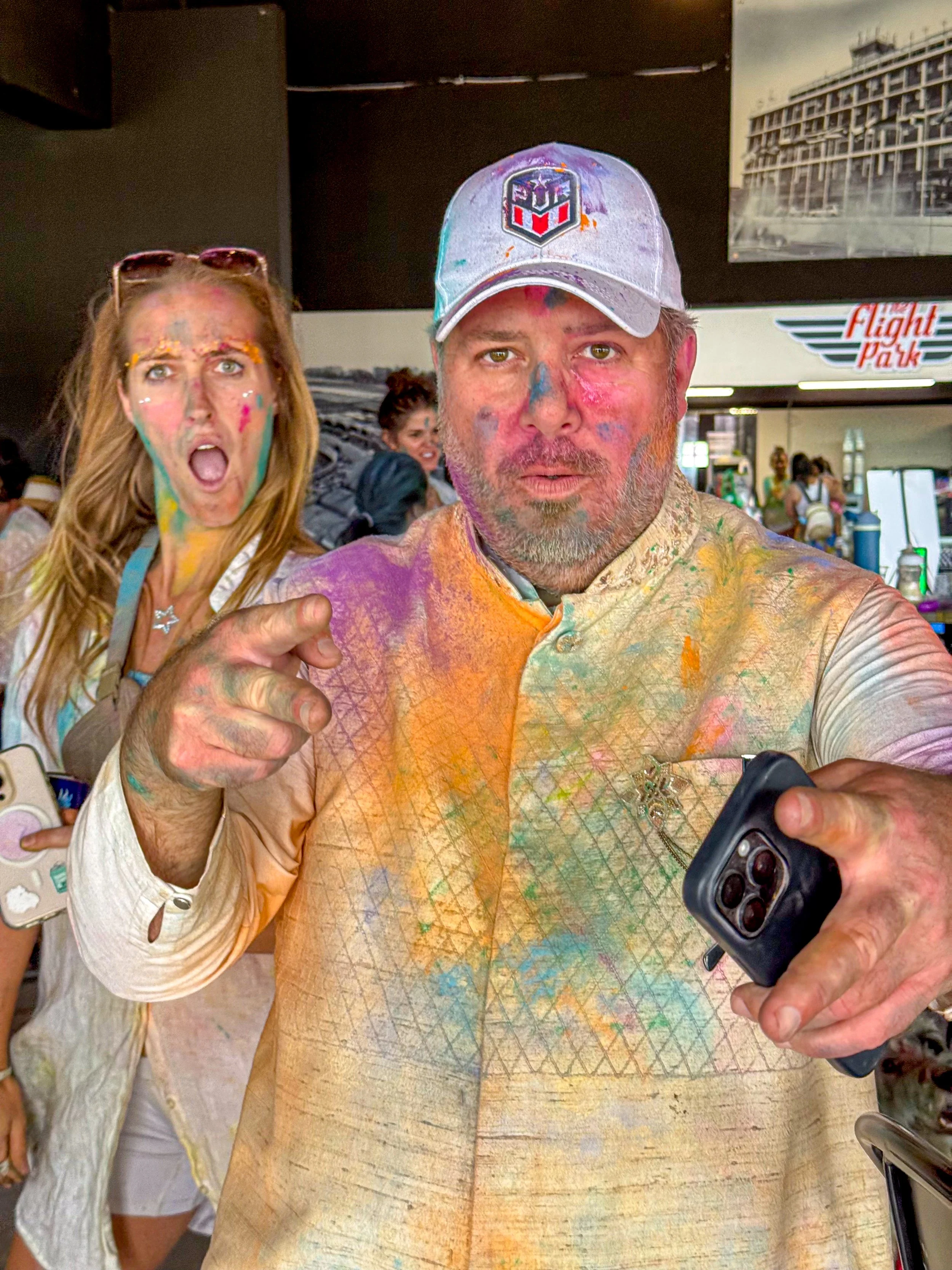 Man and woman covered in colorful powder at a celebration, with the man holding a phone and pointing at the camera; background shows people and an airport terminal sign.