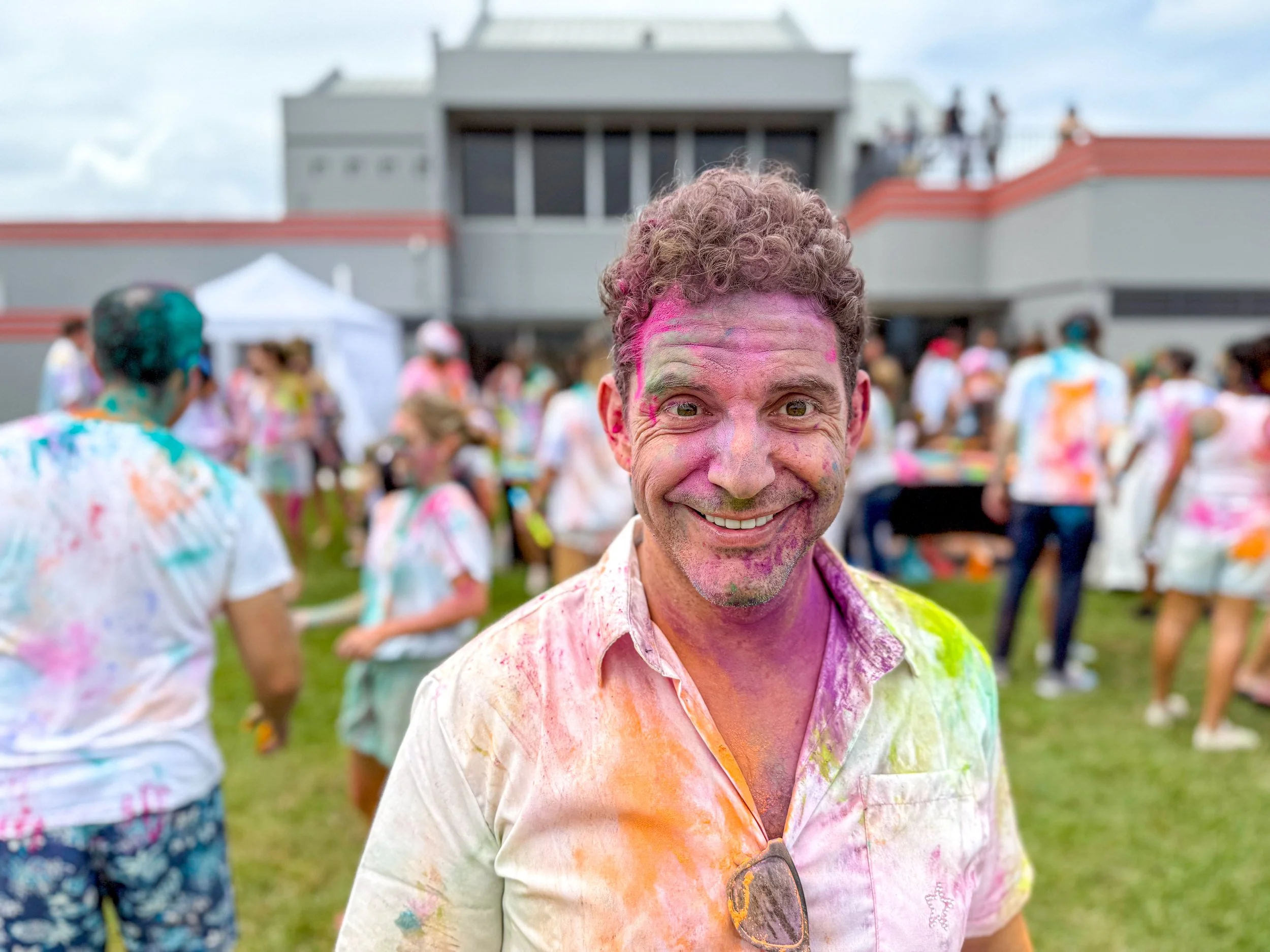 Man smiling at a color run event with colorful powder on his face and shirt, and group of people in the background also covered in colored powder.