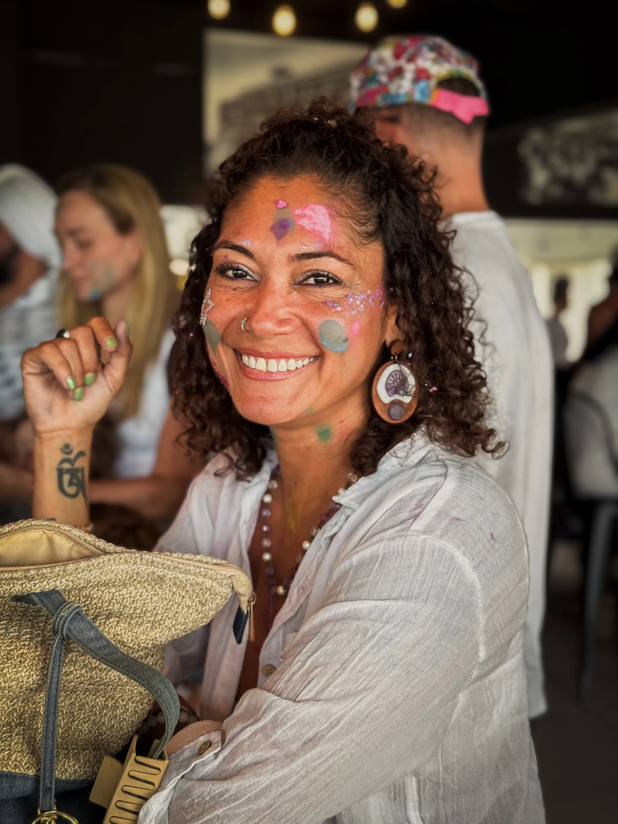 Smiling woman with curly hair, colorful face paint, and large earrings, sitting at a table in a lively indoor setting.