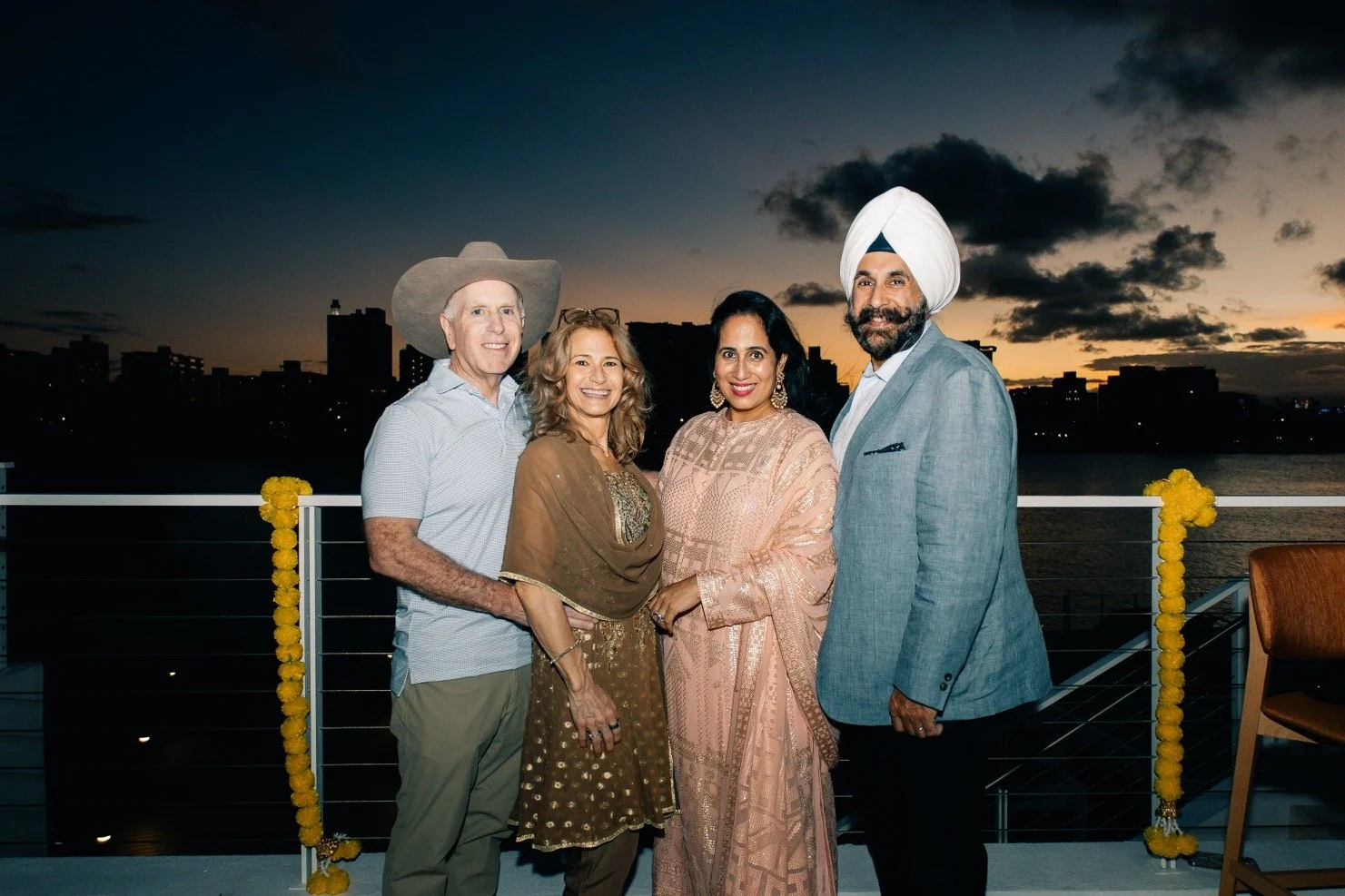 A group of four people, two men and two women, dressed in formal attire, standing on a balcony during sunset with a city skyline in the background. One man is wearing a large hat, and the other man is wearing a turban. They are smiling and posing for
