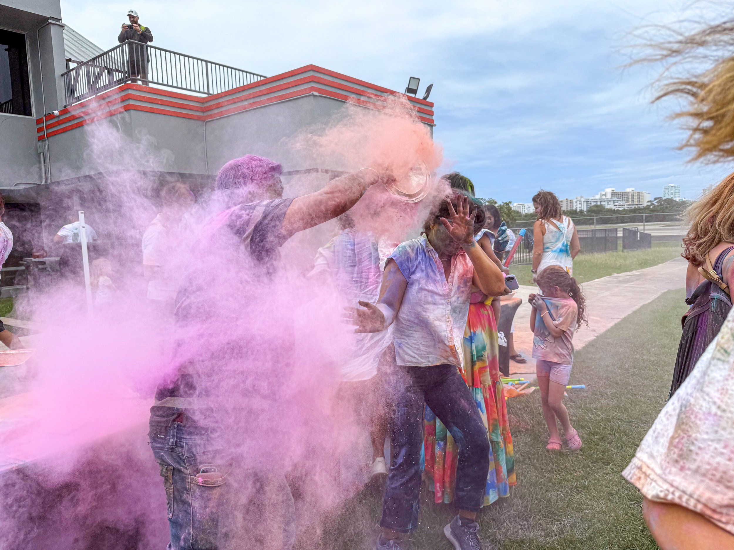 People celebrating Holi with colorful powder on a sunny day, with some individuals throwing pink powder and others capturing the moment.