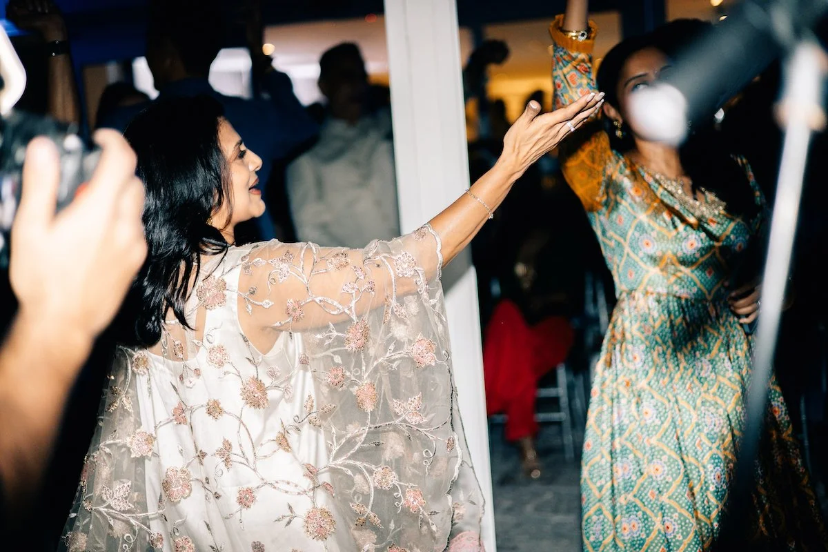 Woman with long black hair wearing a floral embroidered dress dancing at a party with others in the background.