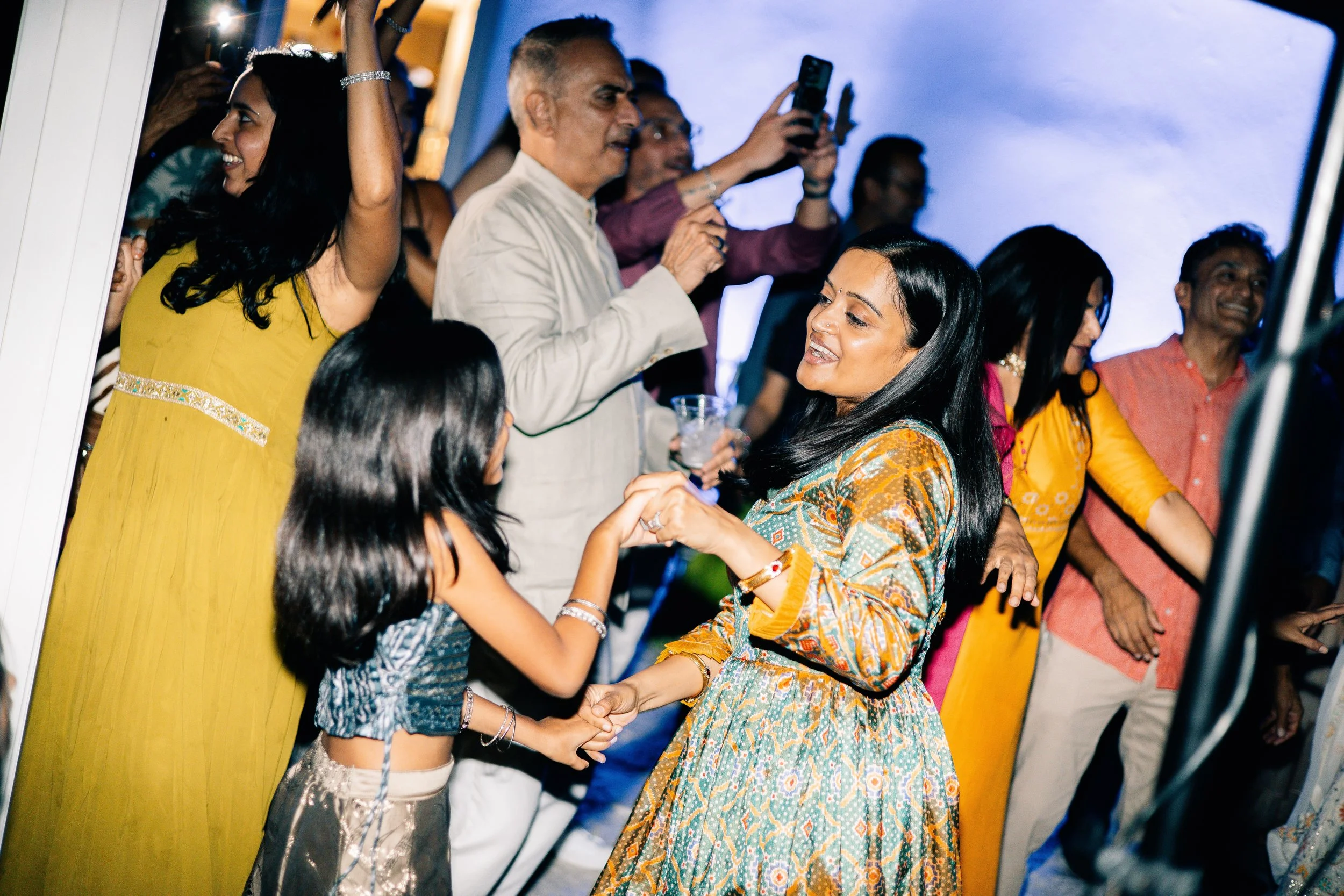 People celebrating at a party, dancing and smiling, with a woman in a colorful dress dancing with a young girl, and other guests in the background.