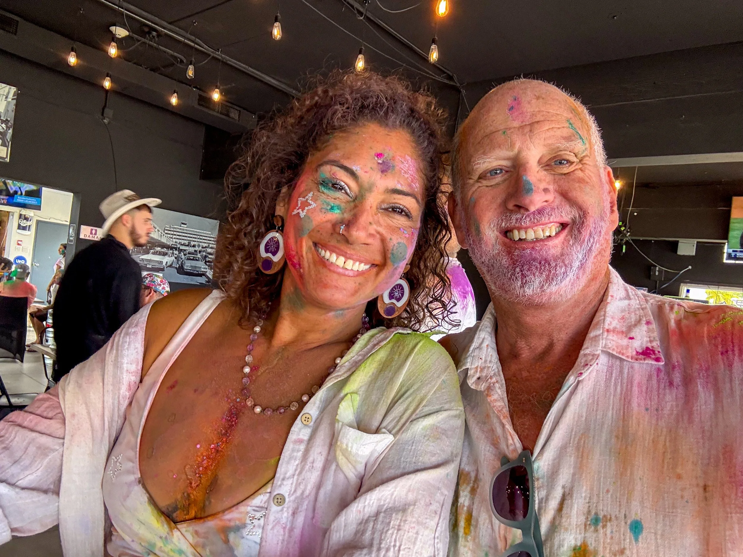 Two smiling people, a woman with curly hair and a man with a beard, covered in colorful paint, celebrating at a color festival or event. They are posing for a selfie in an indoor venue with string lights.
