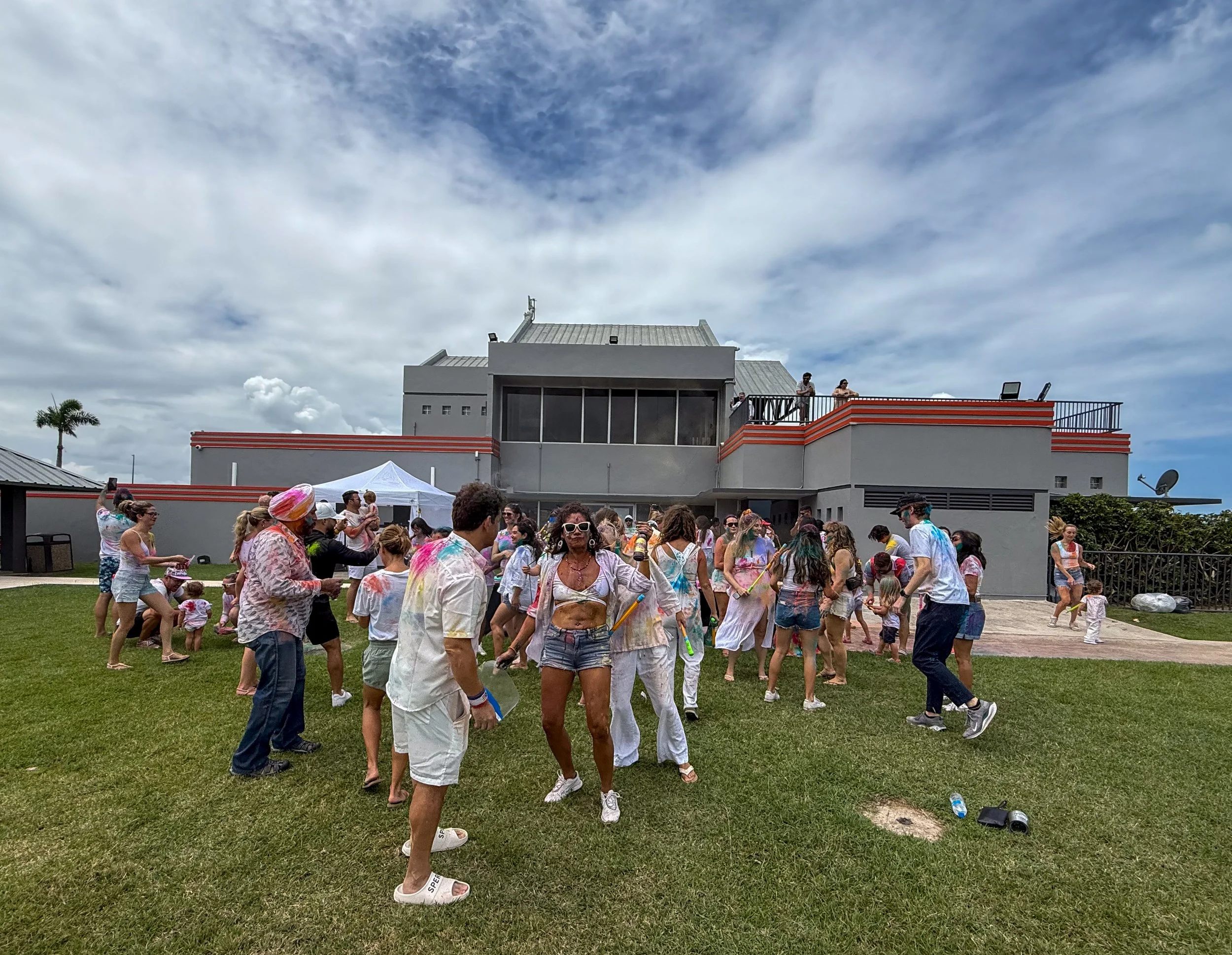 People celebrating outside with colorful powders, dancing and playing on a grassy area near a building under a partly cloudy sky.