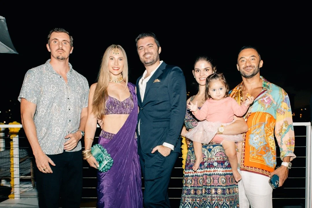 Six people, including a young girl, standing together outdoors at night, dressed in colorful and stylish clothing, smiling and posing for the photo.