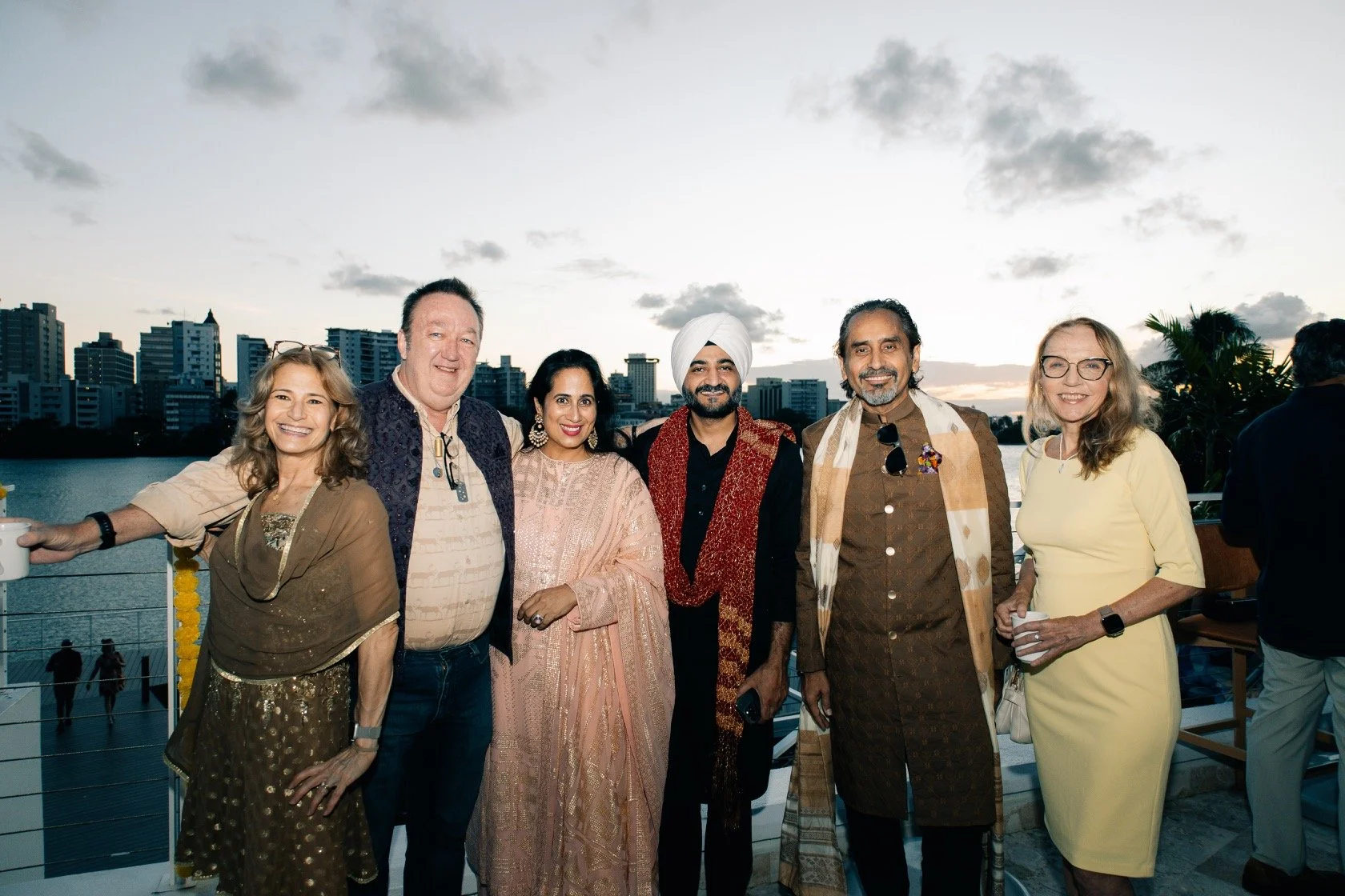 Group of seven diverse people smiling on a terrace overlooking a river with city skyline and cloudy sky at sunset.