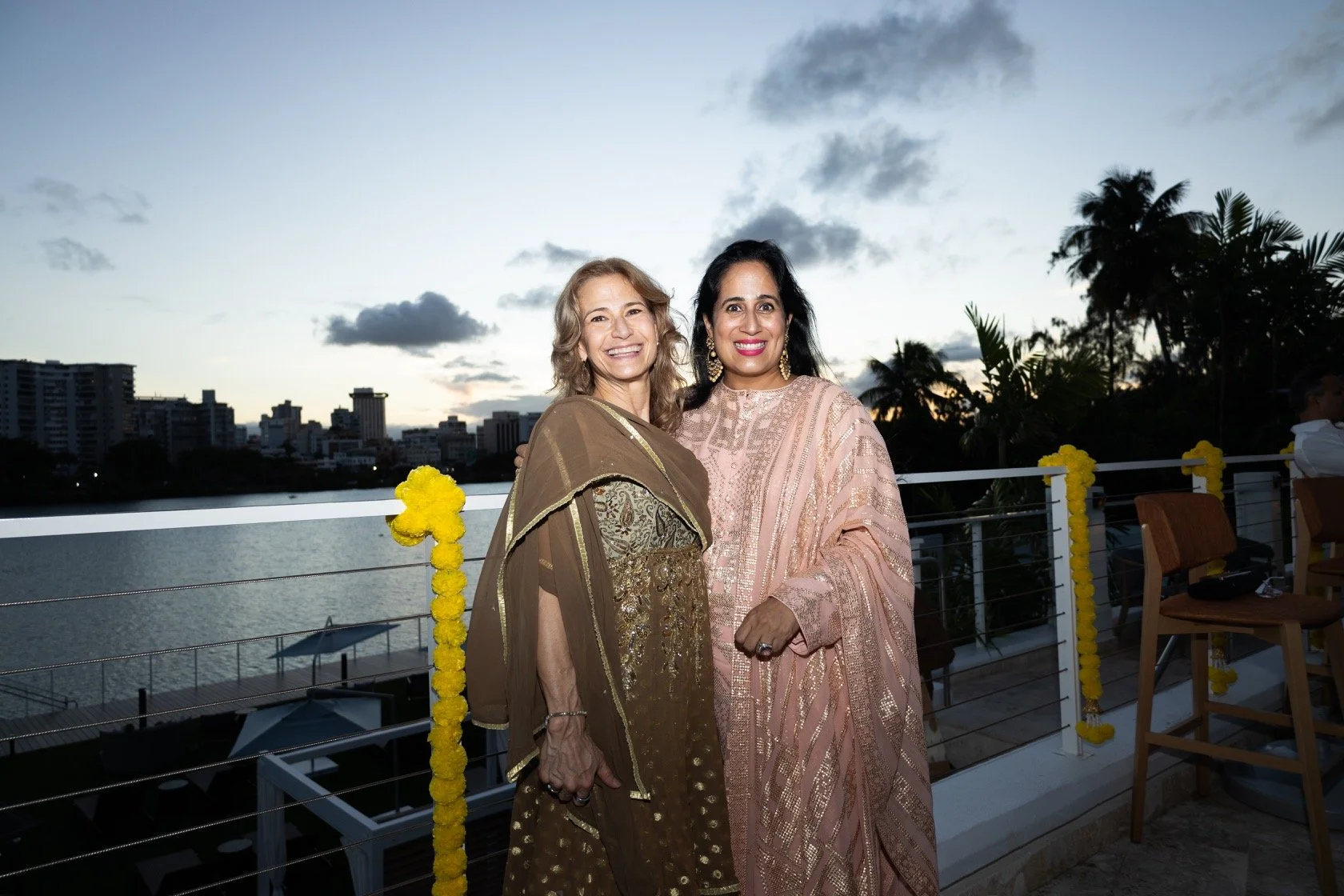 Two women in traditional Indian attire smiling on a balcony during sunset, with city skyline and water in the background.