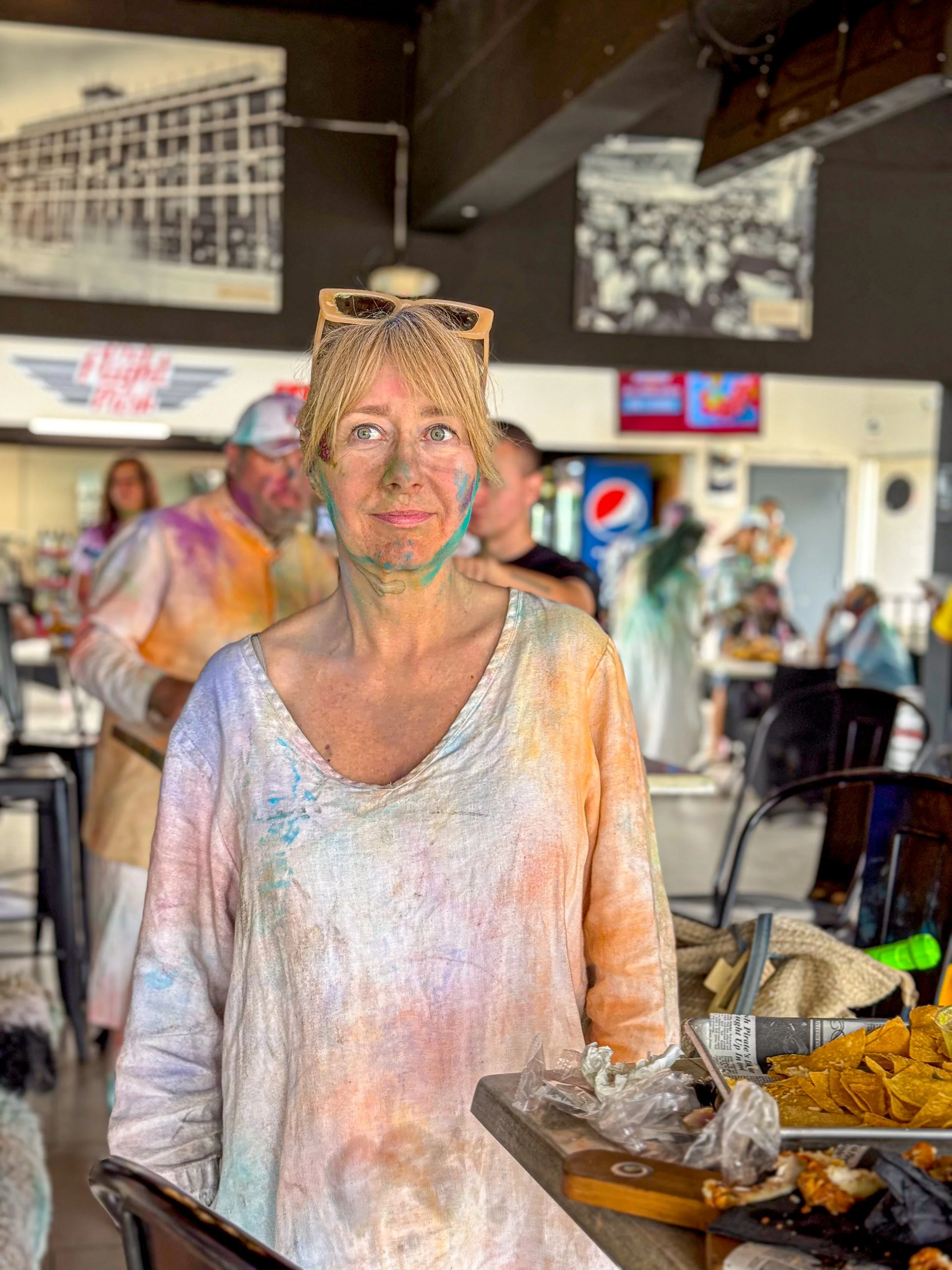 Woman with colorful paint on her face and clothes standing in a restaurant or cafe with people in the background, some also with paint on their faces.