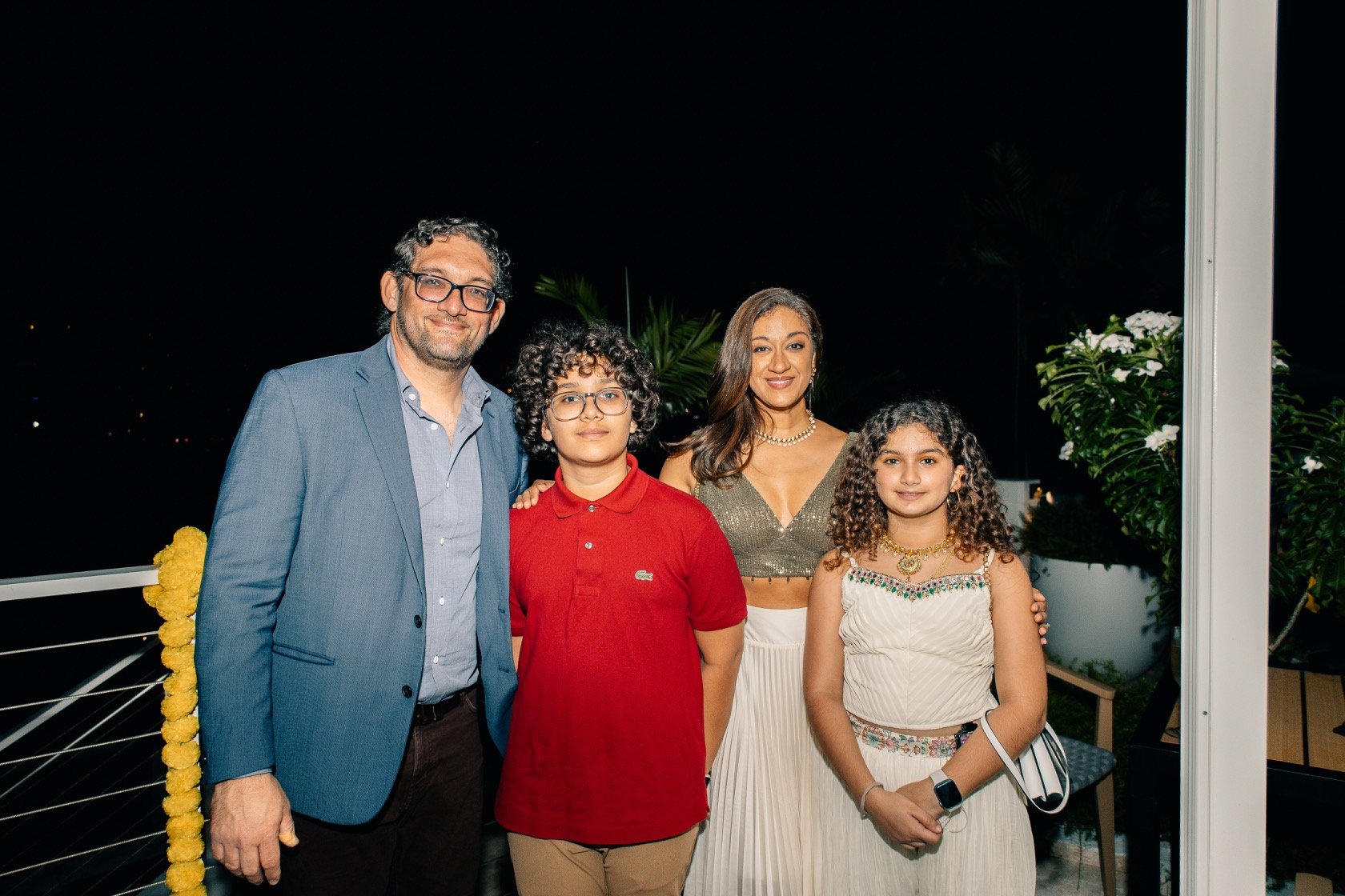 A family of five posing on a balcony at night, with a dark sky and some greenery in the background. The family members are smiling and dressed semi-formally.