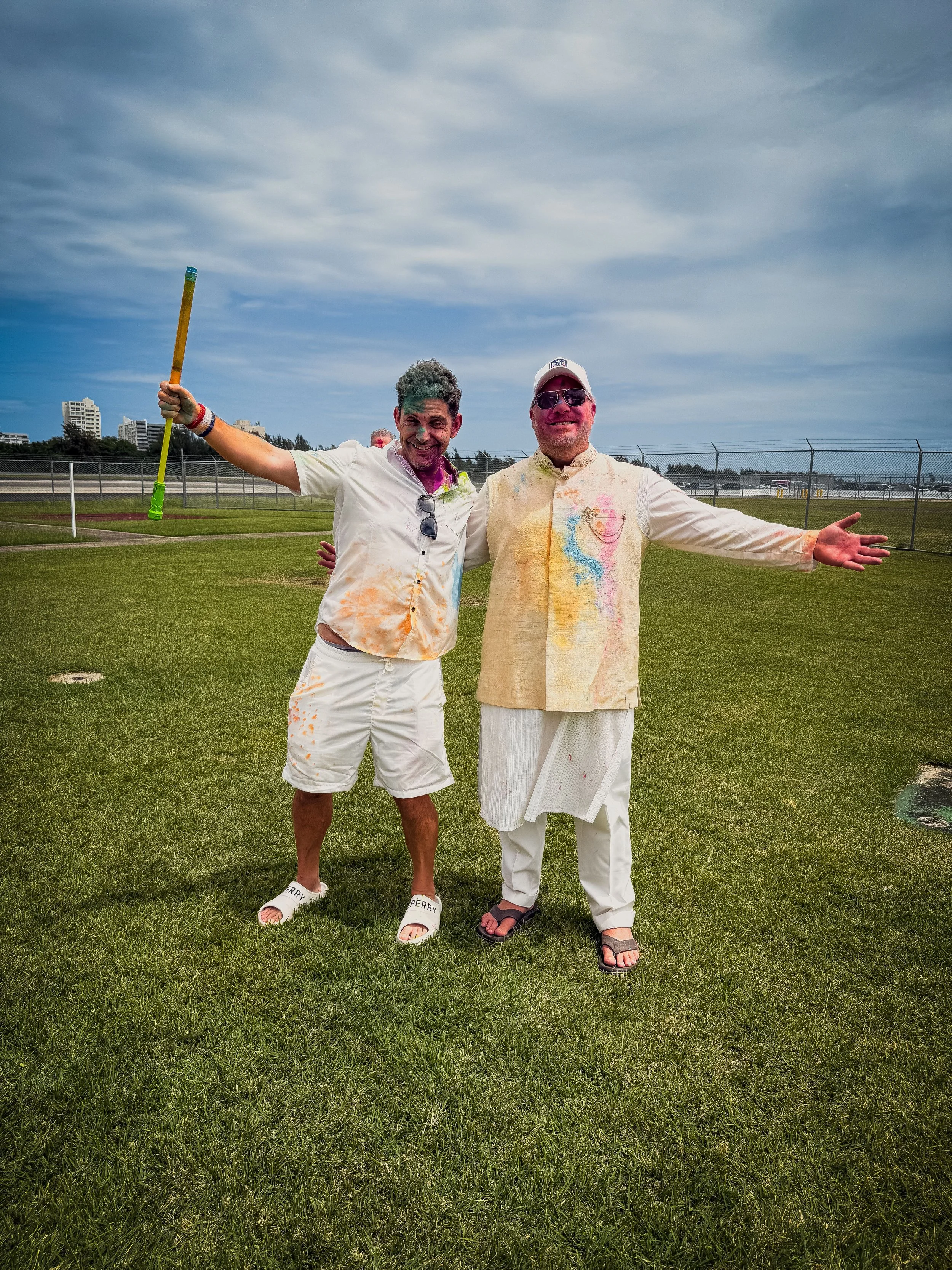Two men celebrating with colorful powder on their clothes and faces, standing on a grassy field under a partly cloudy sky, with buildings in the distance.