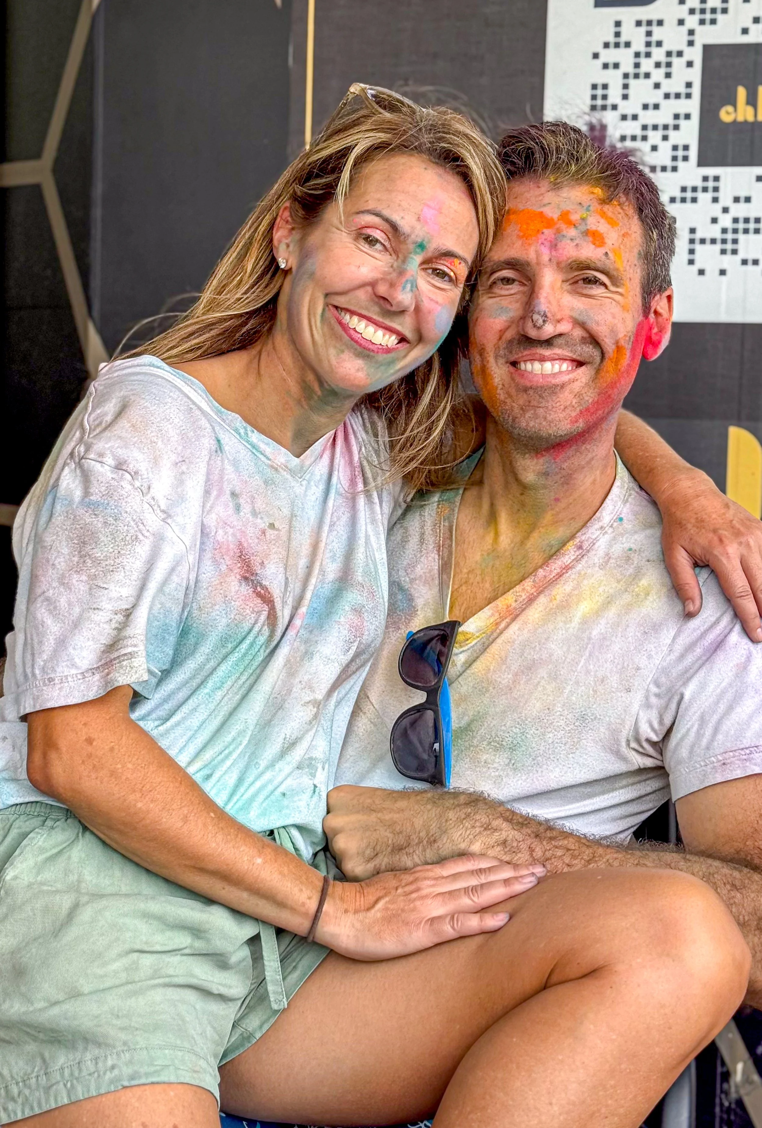 Smiling woman and man with colorful powder smeared on their faces and clothes, sitting closely together after a festival or celebration.