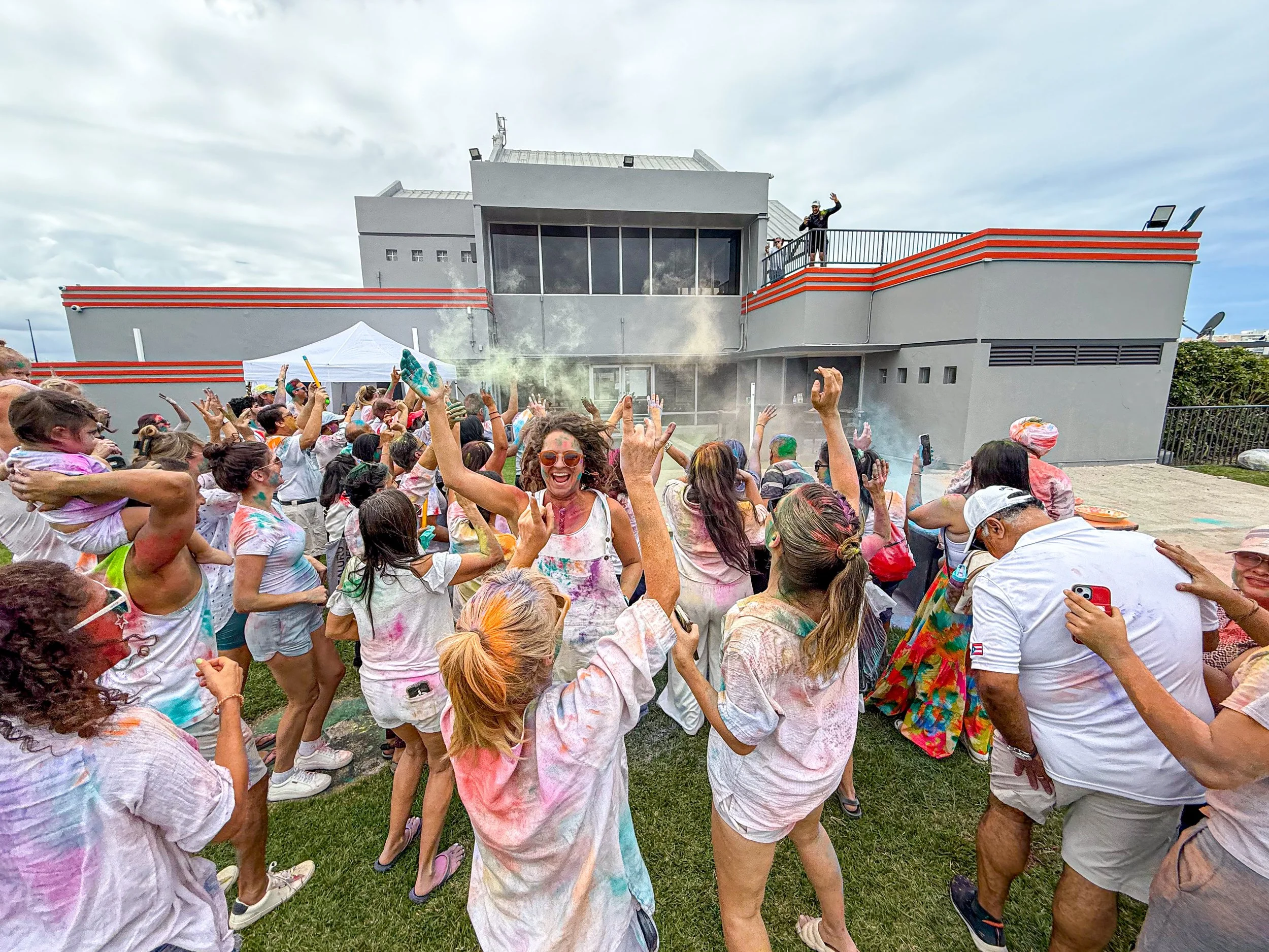 People celebrating a color run event with powder on their clothes and faces, outdoors near a grey building, with some on a balcony and others dancing on the grass.