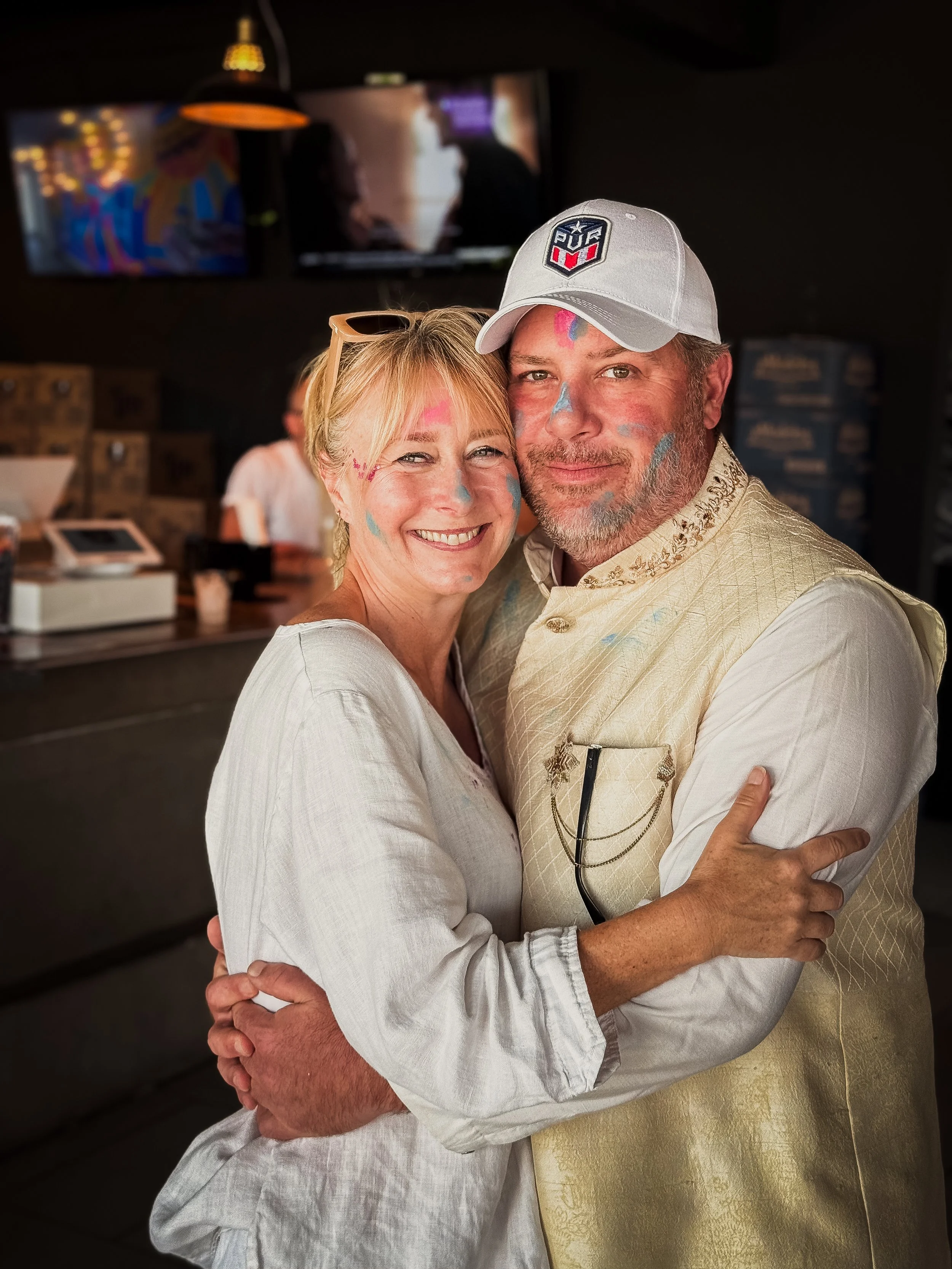A smiling woman and a man are hugging and looking at the camera, with colorful paint on their faces. The woman has sunglasses on her head, and the man is wearing a white cap and a cream-colored vest. They are in a dimly lit indoor setting with televi