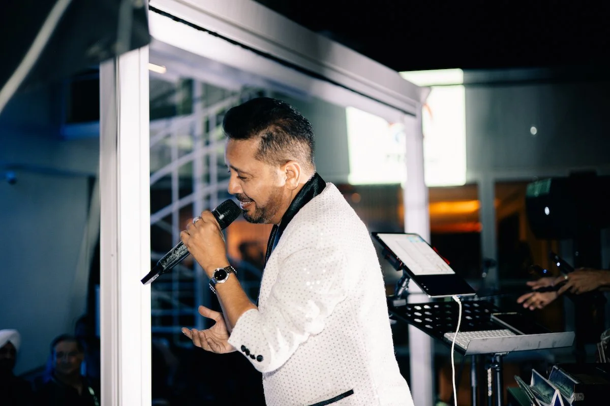 A man wearing a white suit jacket singing into a microphone at an event.
