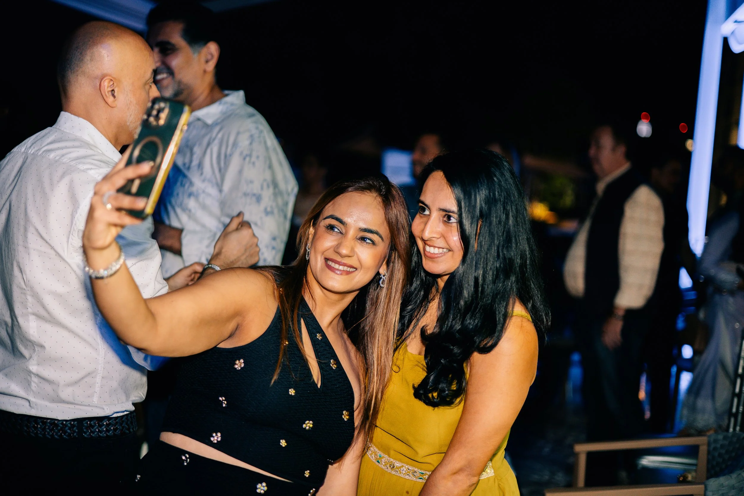 Two women taking a selfie together at a nighttime event, surrounded by other people socializing.