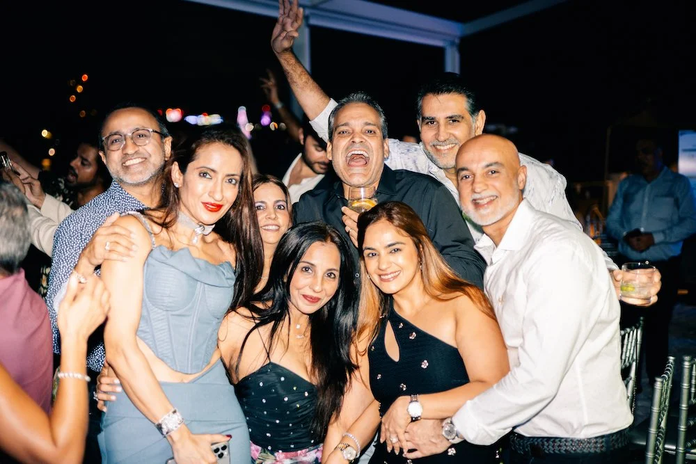 Group of people celebrating at a party, smiling and holding drinks, at night with colorful lights in the background.