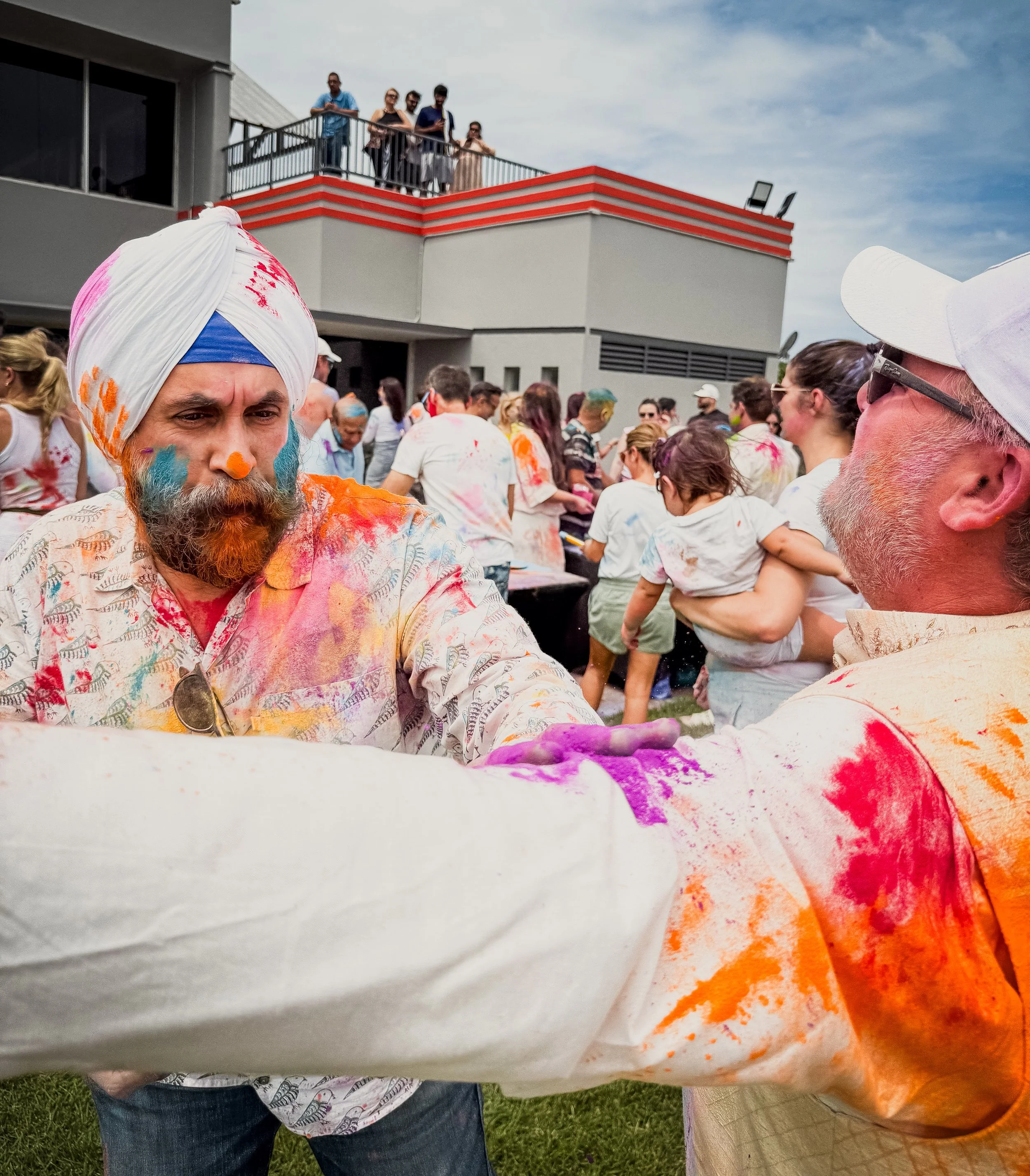 Two men with colorful powder on their faces and clothes participate in a color festival, surrounded by a crowd of people also covered in colored powder outdoors near a modern building.