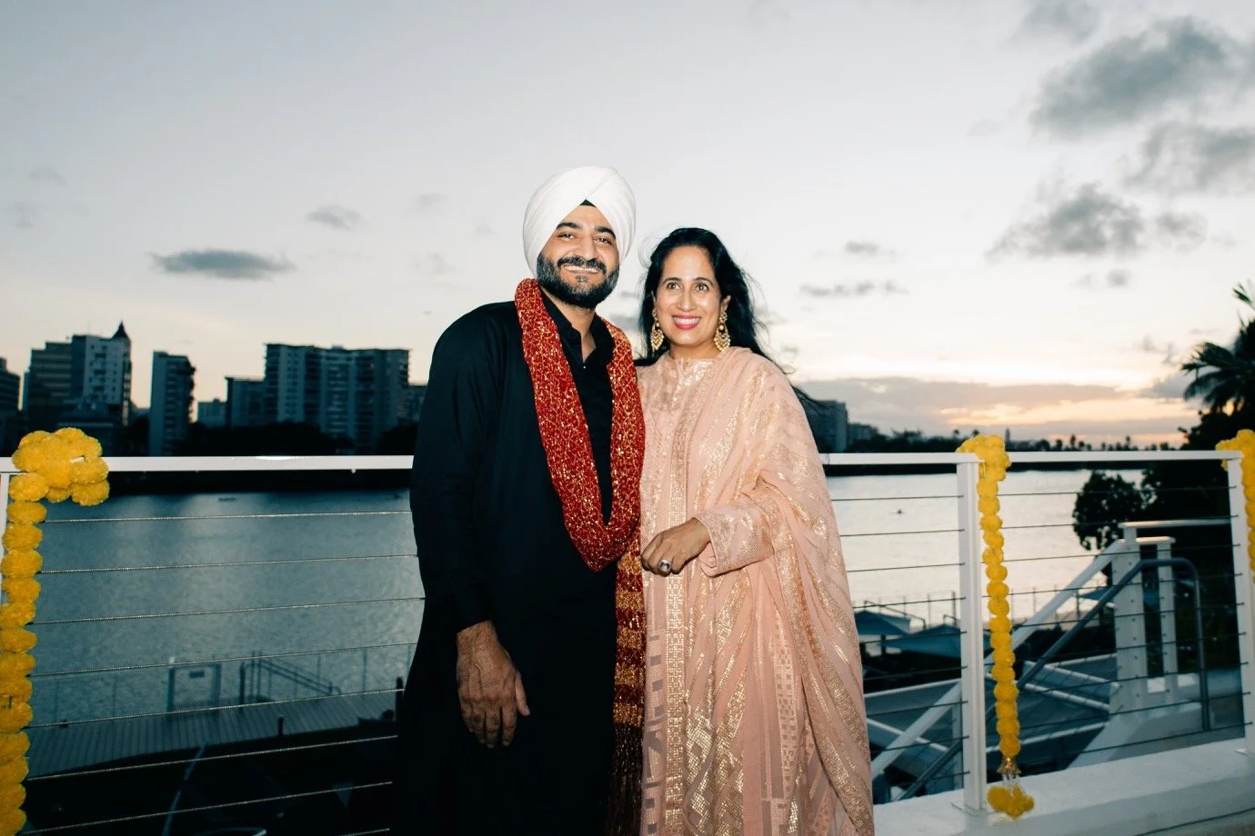 A man in traditional Indian attire with a white turban and a woman in a pink saree posing together on a balcony during sunset, with city buildings in the background.