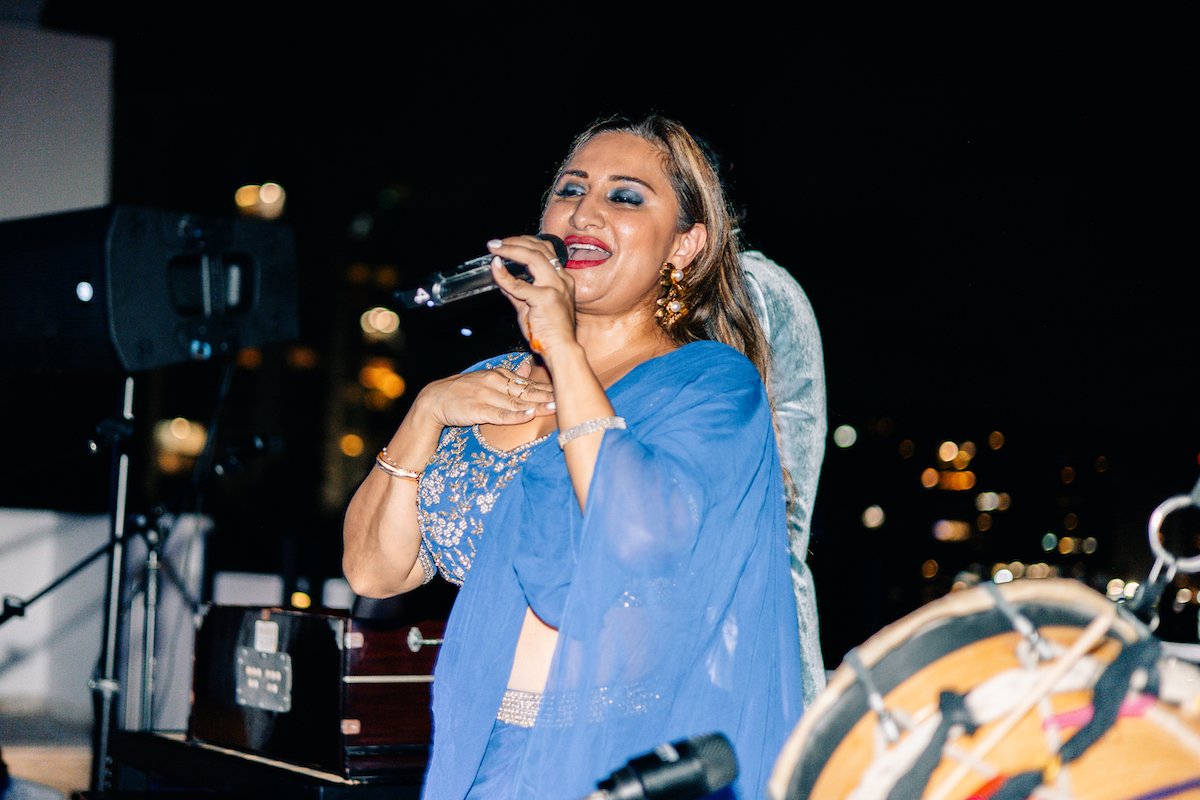 A woman singing into a microphone at night, wearing a blue dress with silver embroidery, gold earrings, and makeup, with city lights in the background.
